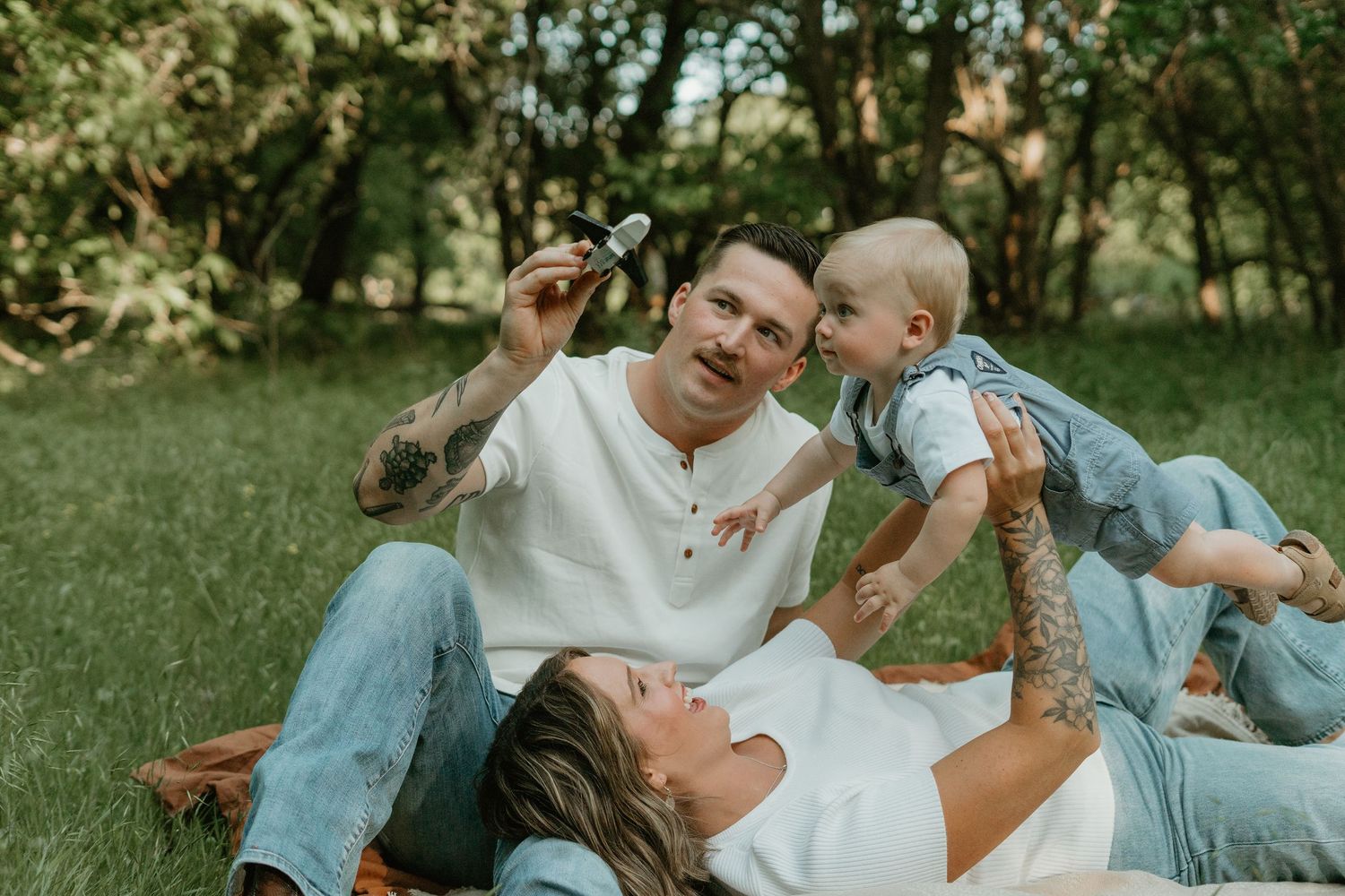 Happy family sits on picnic blanket in park, father holds flower while mother cradles laughing baby.