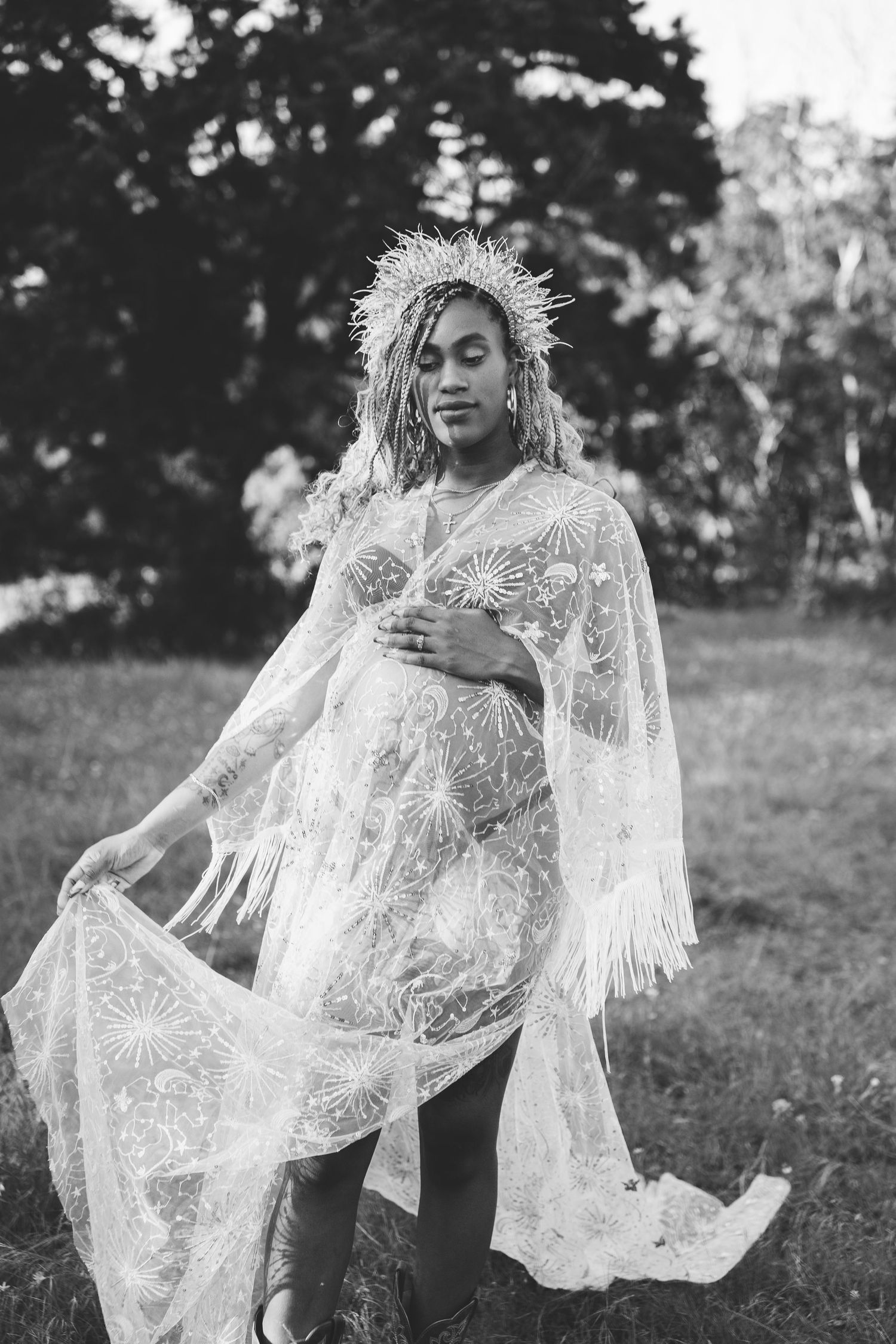 Black and white photo of a pregnant woman in a fringed lace dress and crown in a field.
