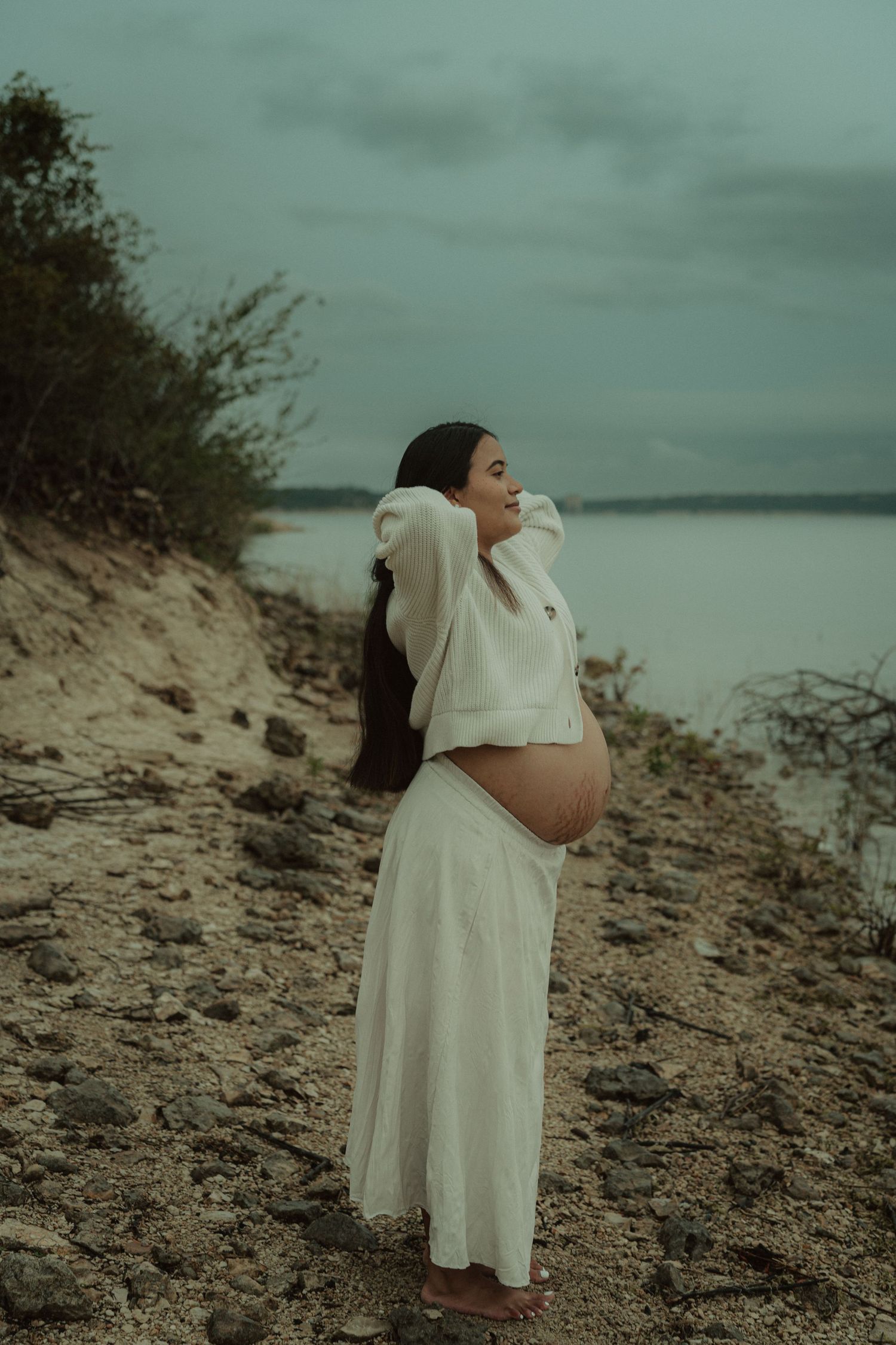 A pregnant woman in a white dress and sweater stands on a rocky beach under cloudy skies.