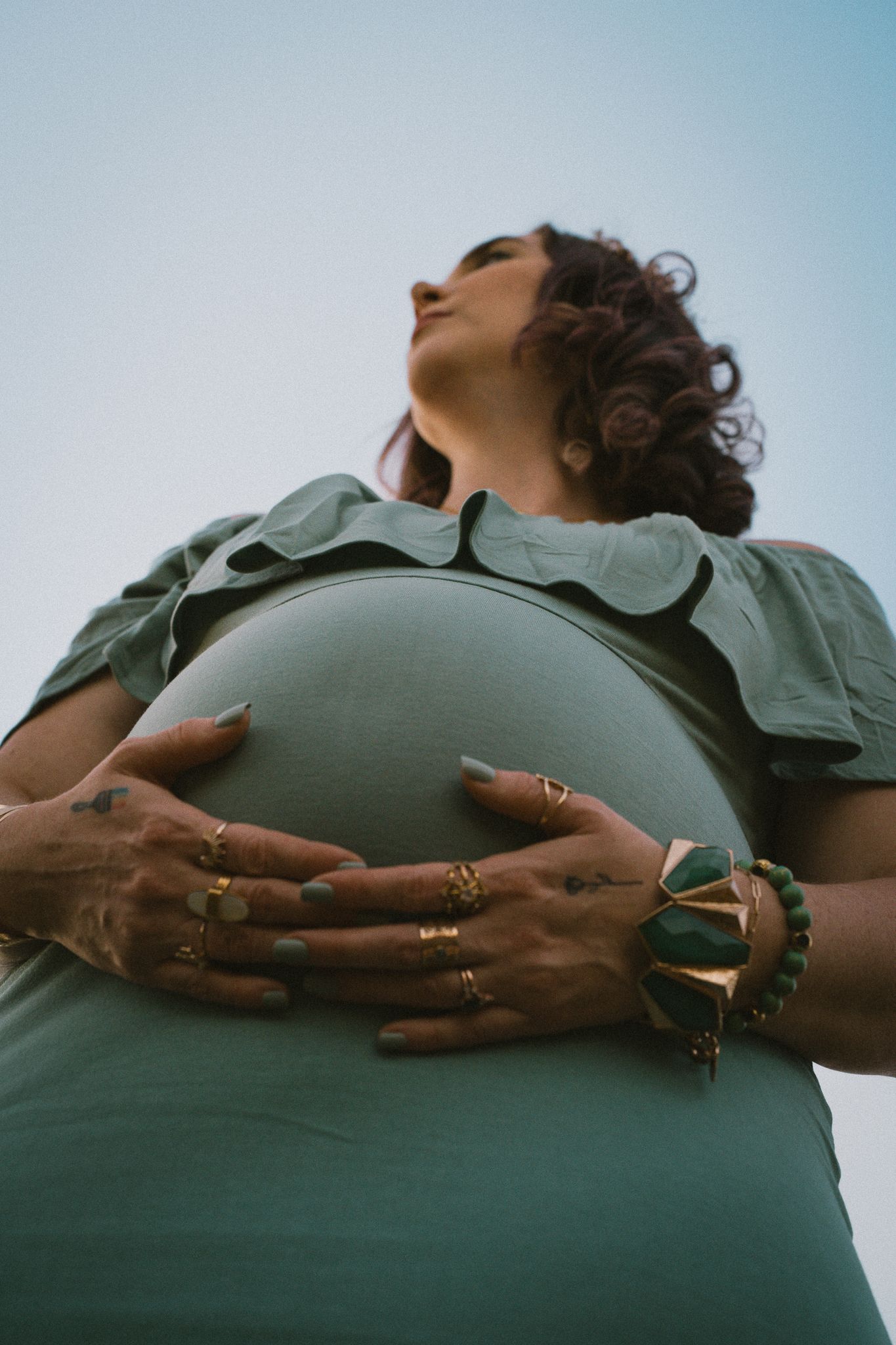 Low angle view of pregnant woman in sage green dress cradling baby bump against blue sky.