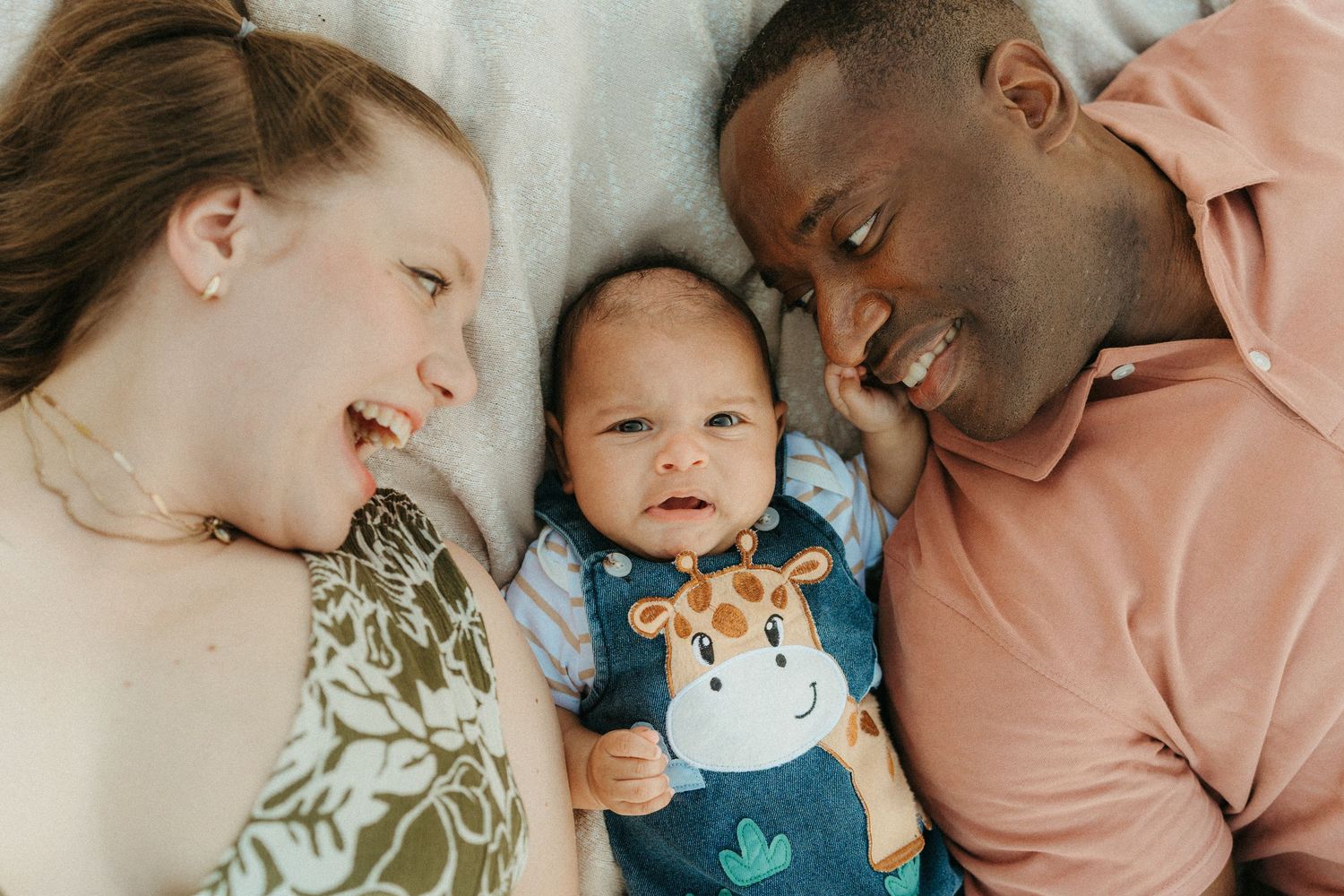 Interracial couple lying down smiling at their newborn baby dressed in a giraffe onesie.