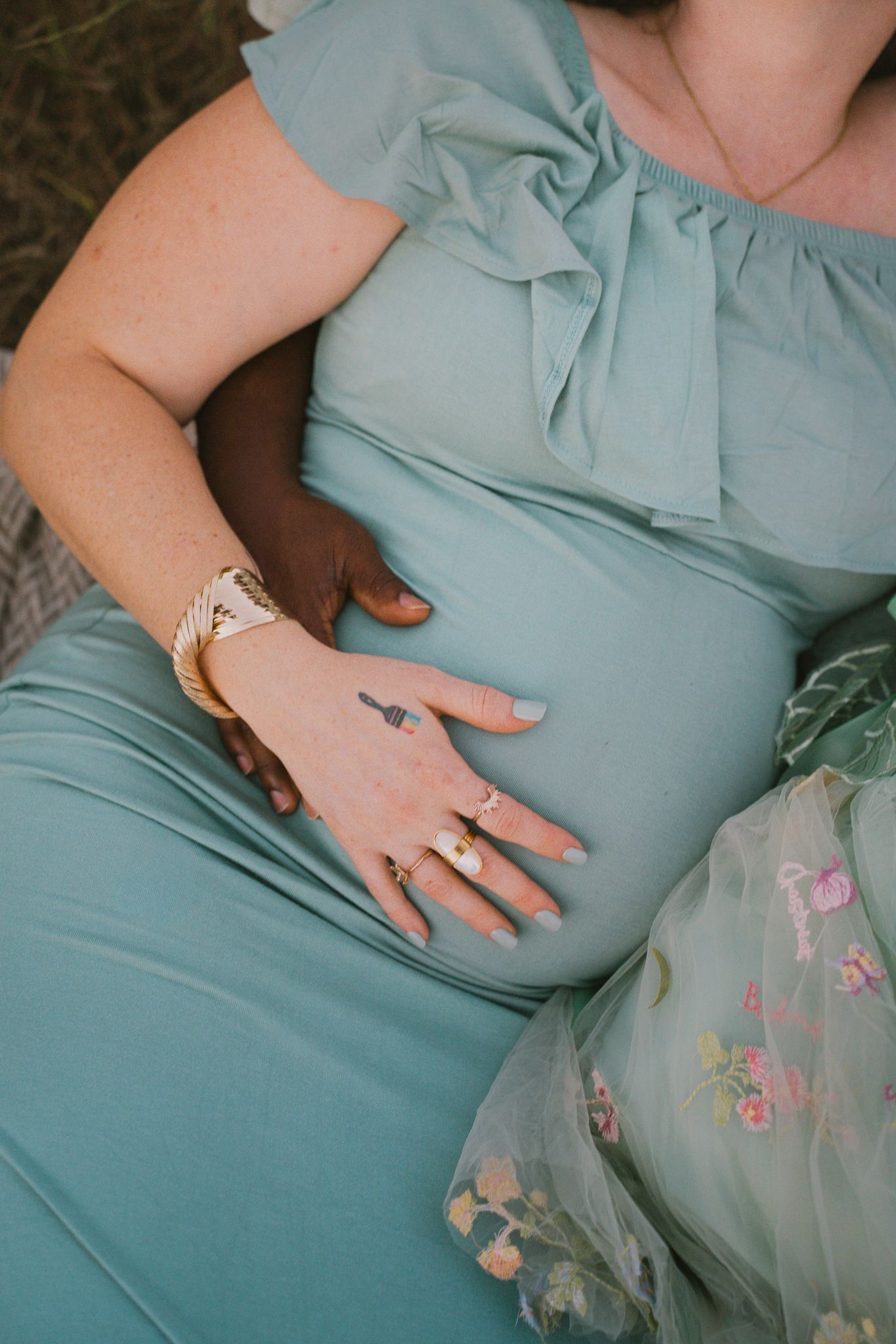 Close-up of interracial couple's hands on pregnant belly in sage green maternity dress.