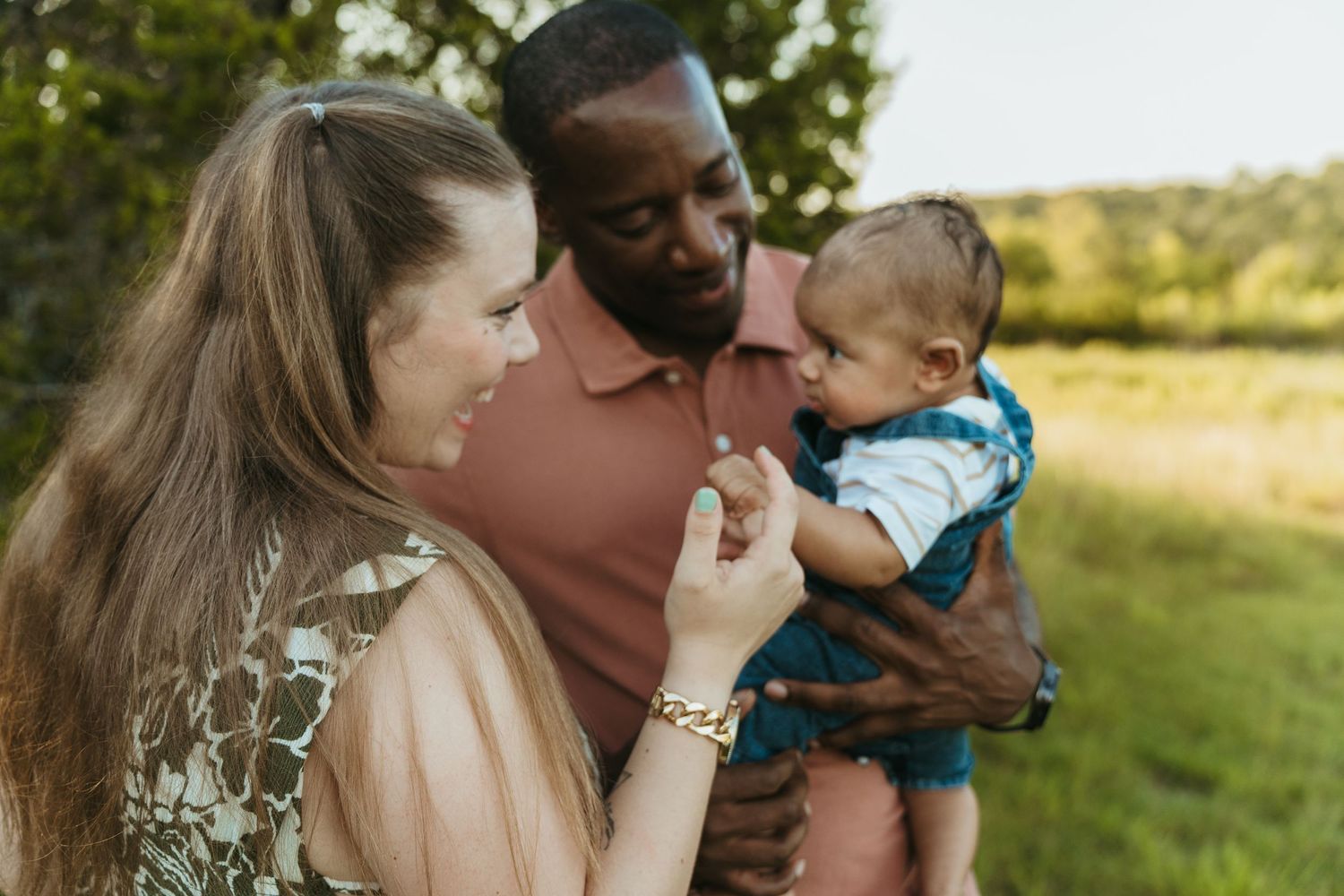 Smiling interracial couple standing outdoors holding their infant baby in a sunny park.