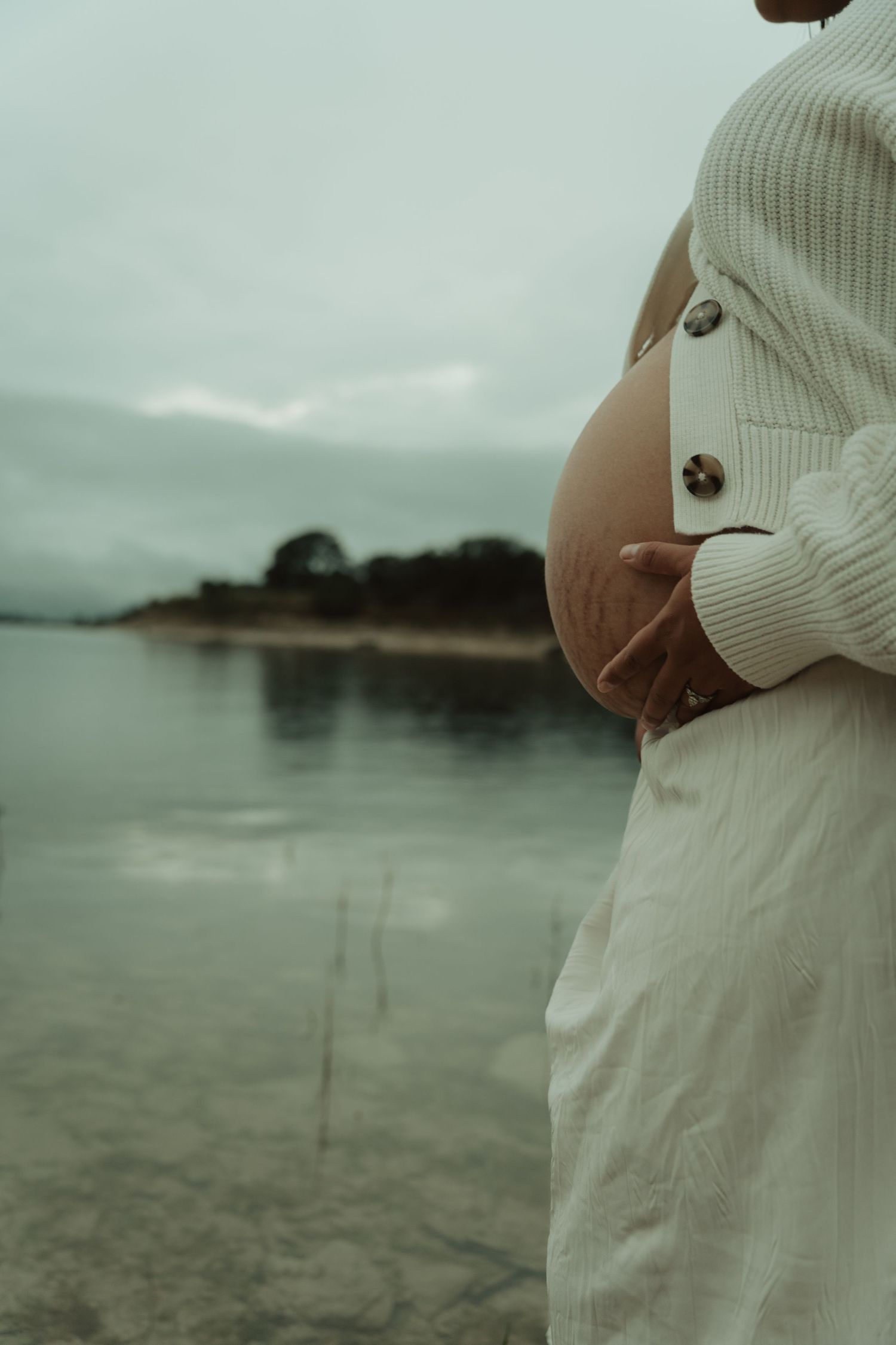 A close-up of a pregnant woman in a white outfit cradling her bare baby bump by a lake.