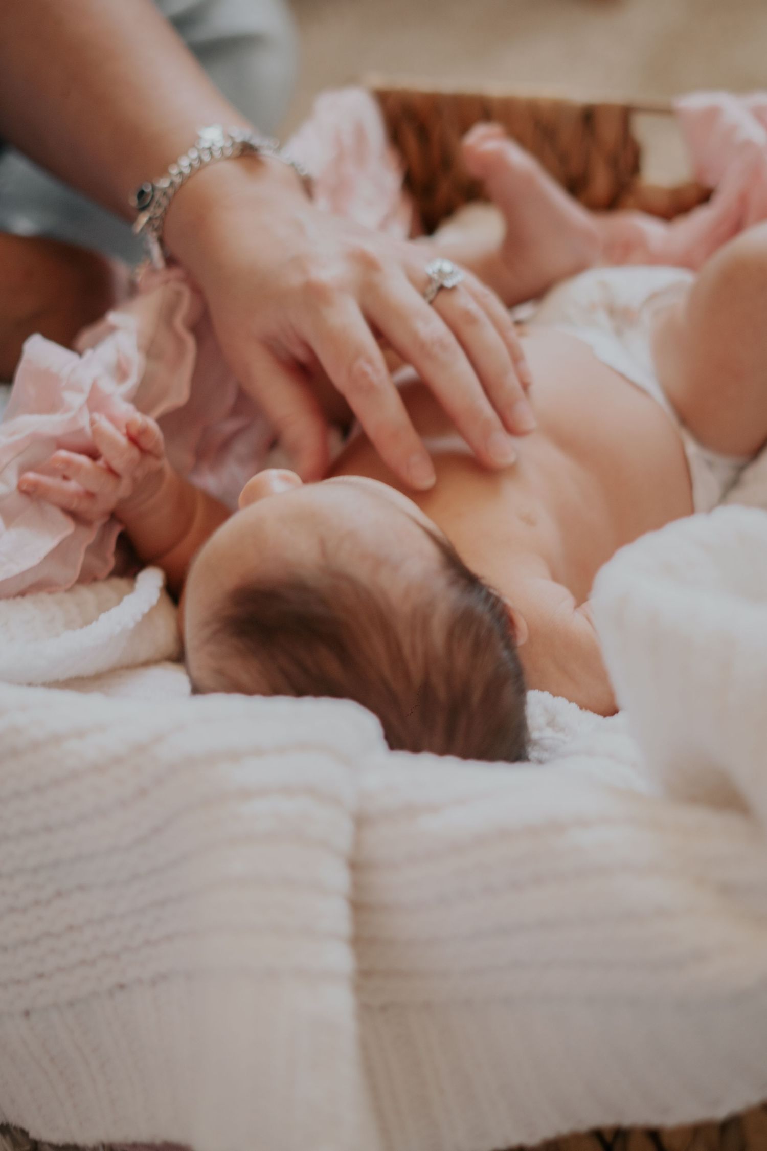 Adult hands gently touching a sleeping newborn baby lying on a soft white knit blanket in a wooden basket.