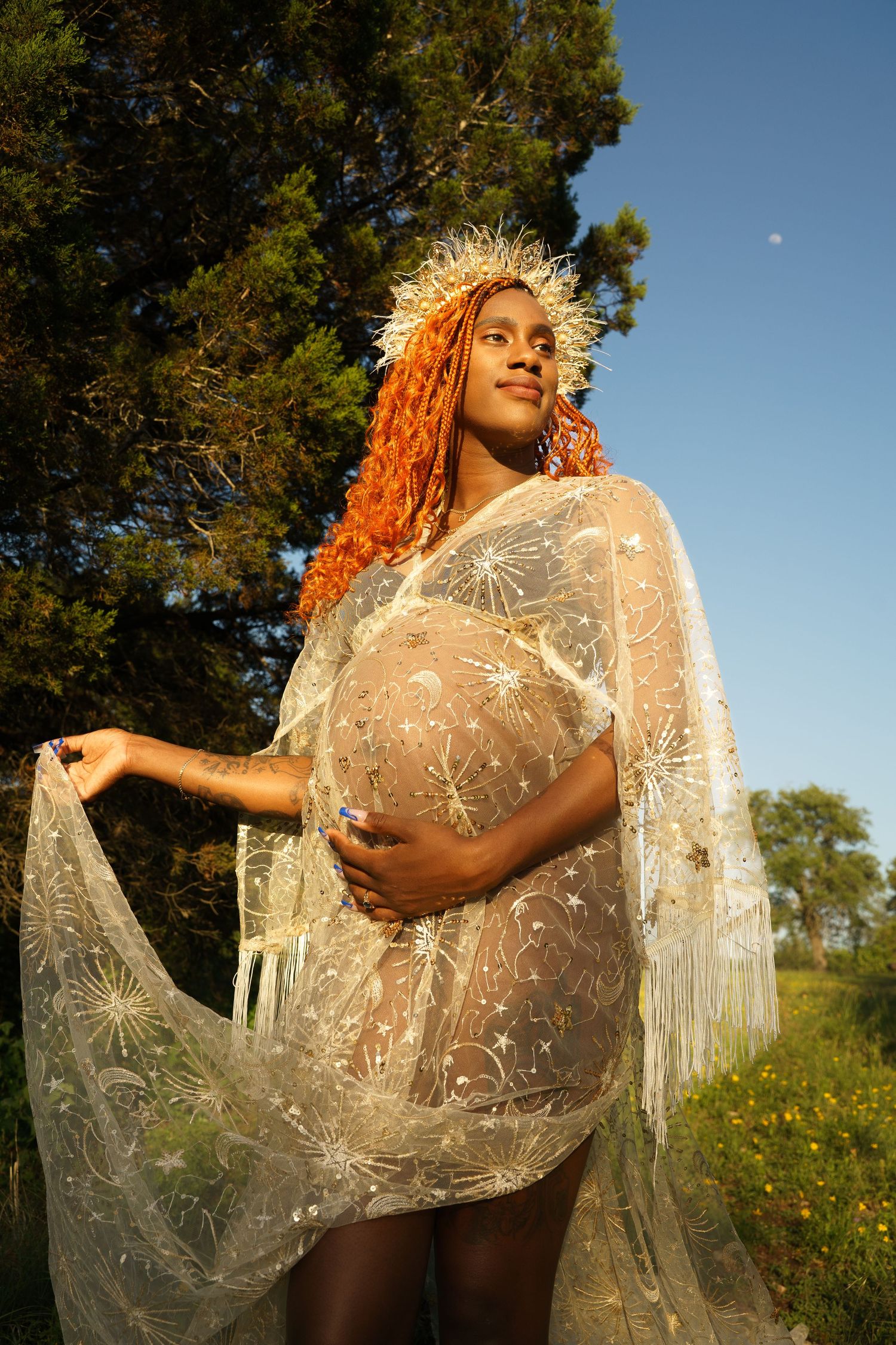 Pregnant woman with orange braids in a sheer lace dress and crown posing in golden sunlight.