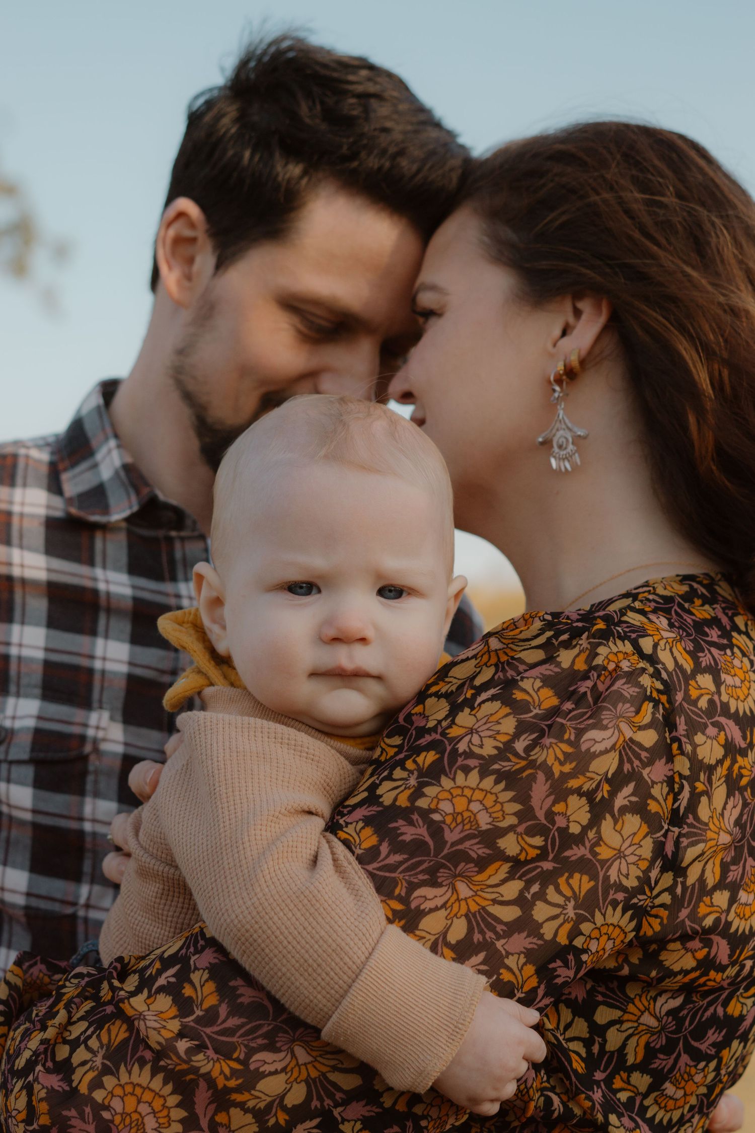 Loving parents press foreheads together while holding their alert baby in warm golden light.