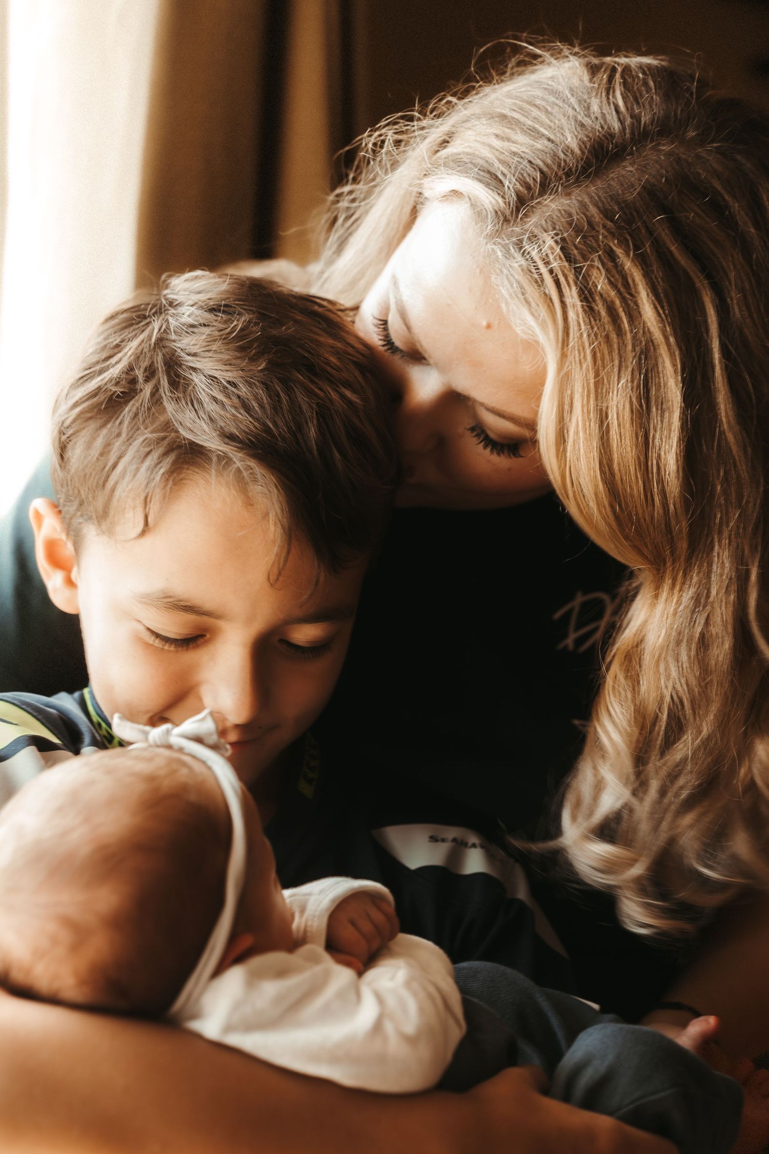 A mother with blonde hair kisses her young son as they both gaze at a newborn baby.