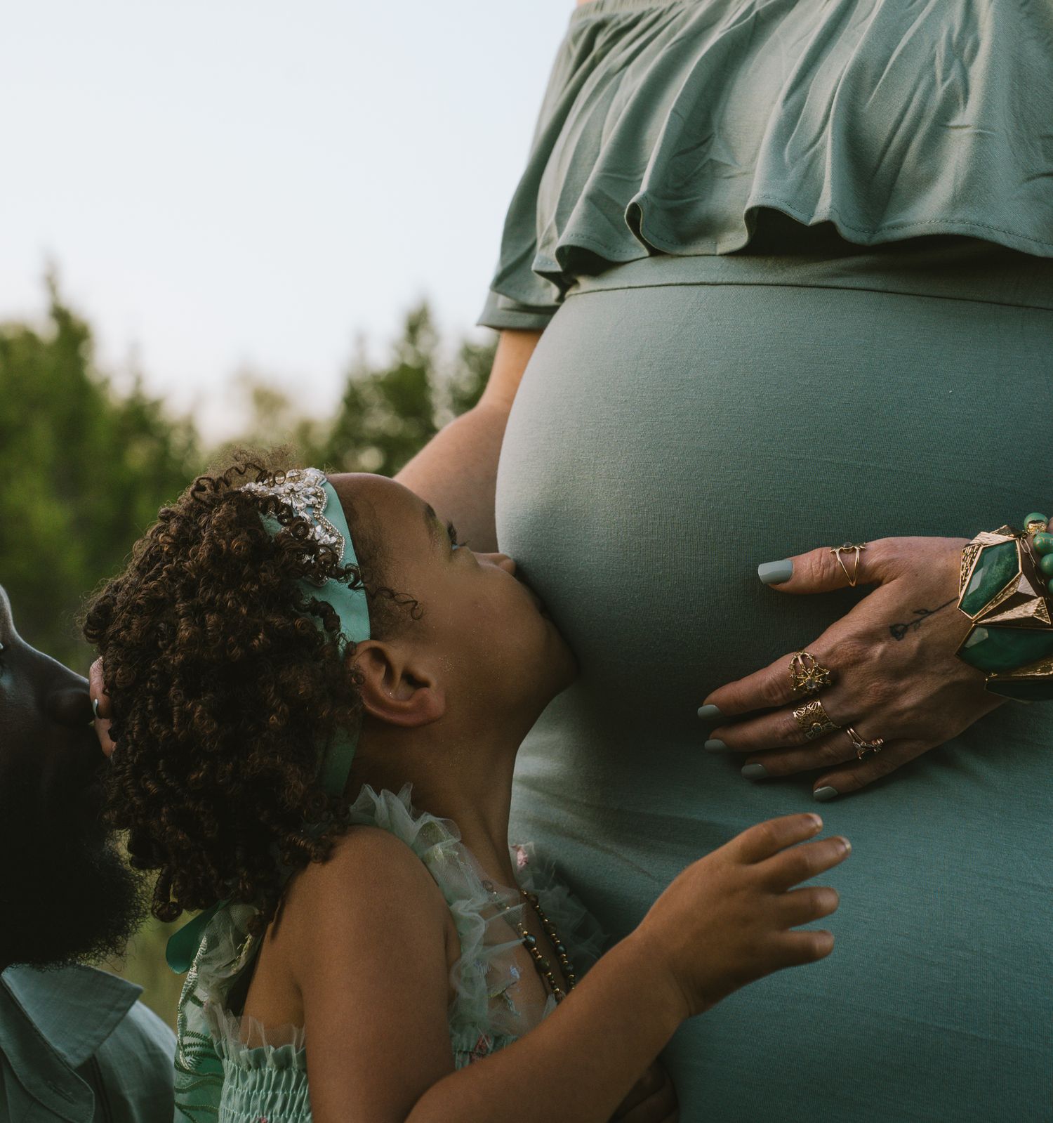 A young girl kisses her mother's pregnant belly during a maternity photo shoot outdoors.