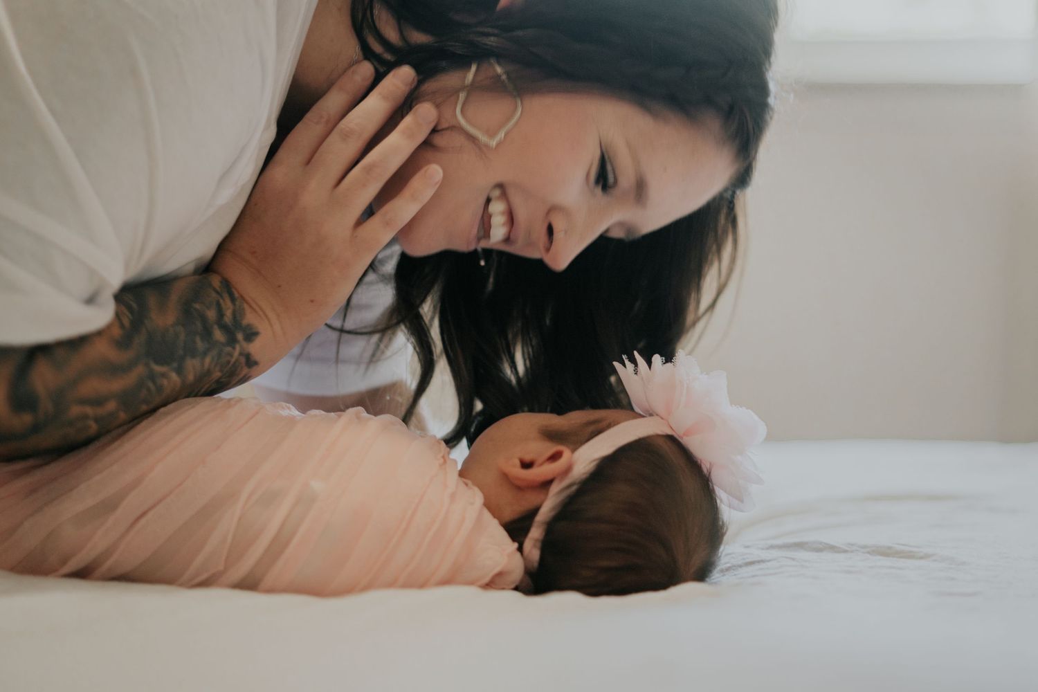 Smiling tattooed mother leaning over her newborn baby lying on a white bed wearing a pink headband.