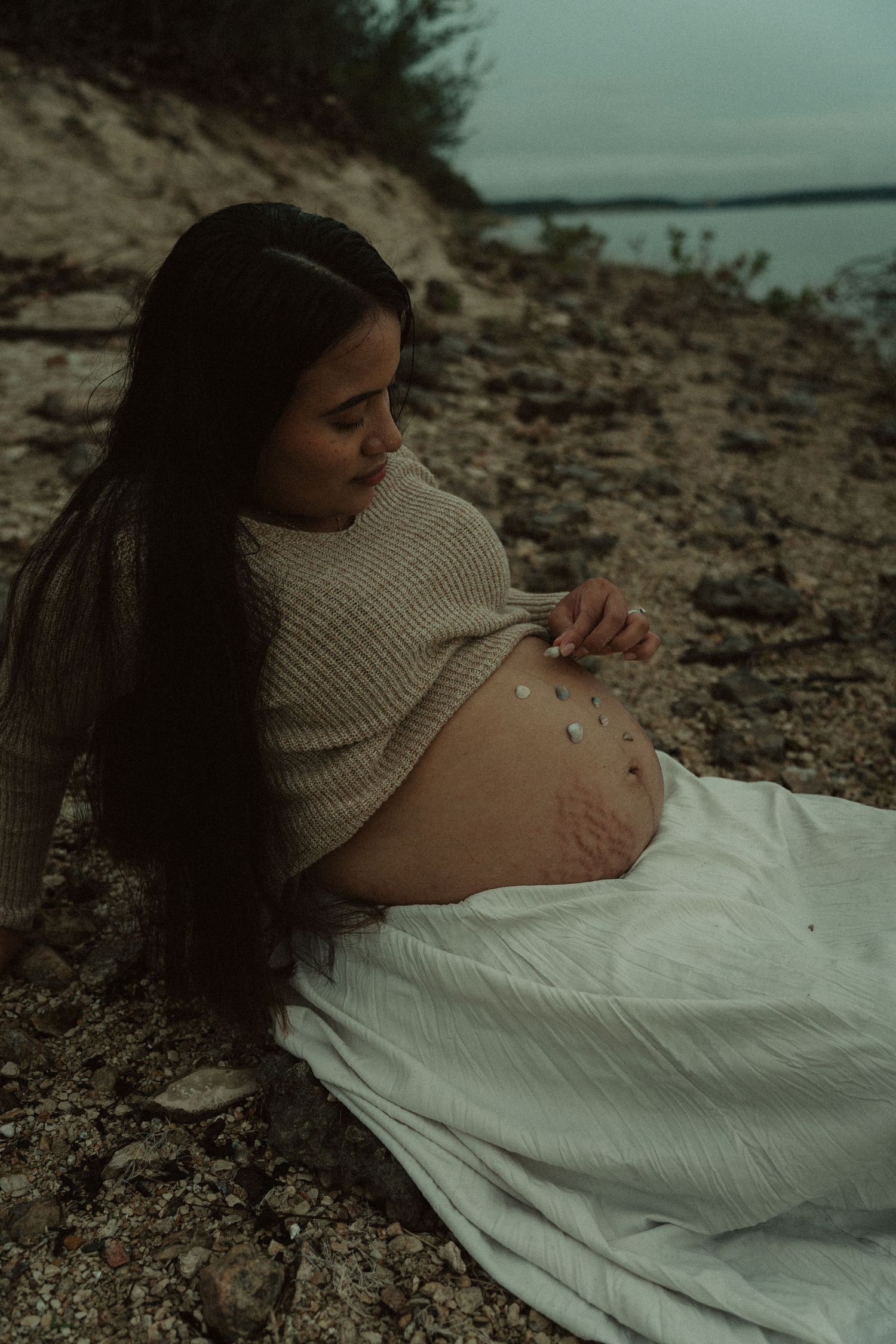 A pregnant woman in a beige sweater sits on a rocky shore placing stones on her belly.