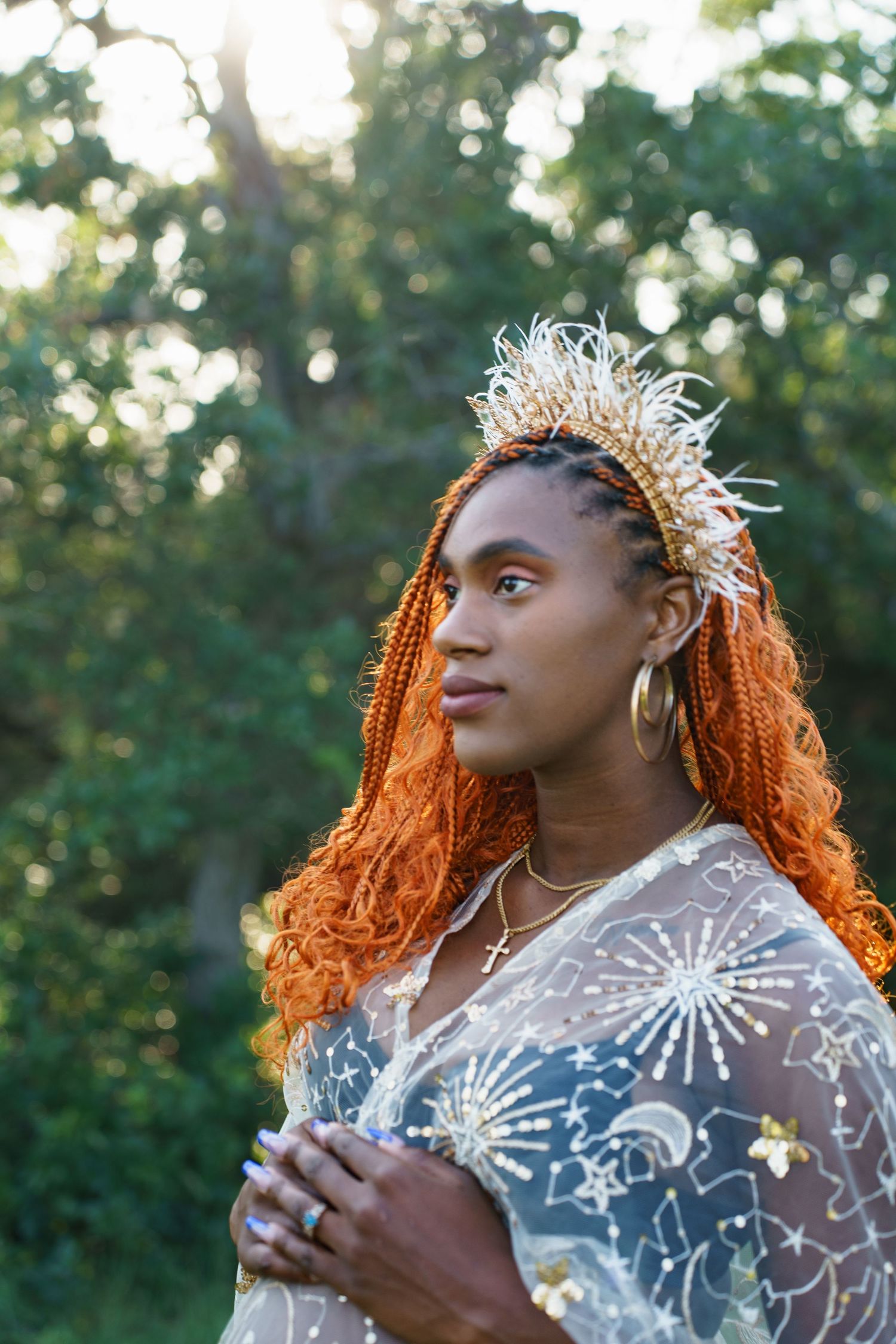 Black woman with orange braids wearing a decorative crown and celestial lace dress outdoors.