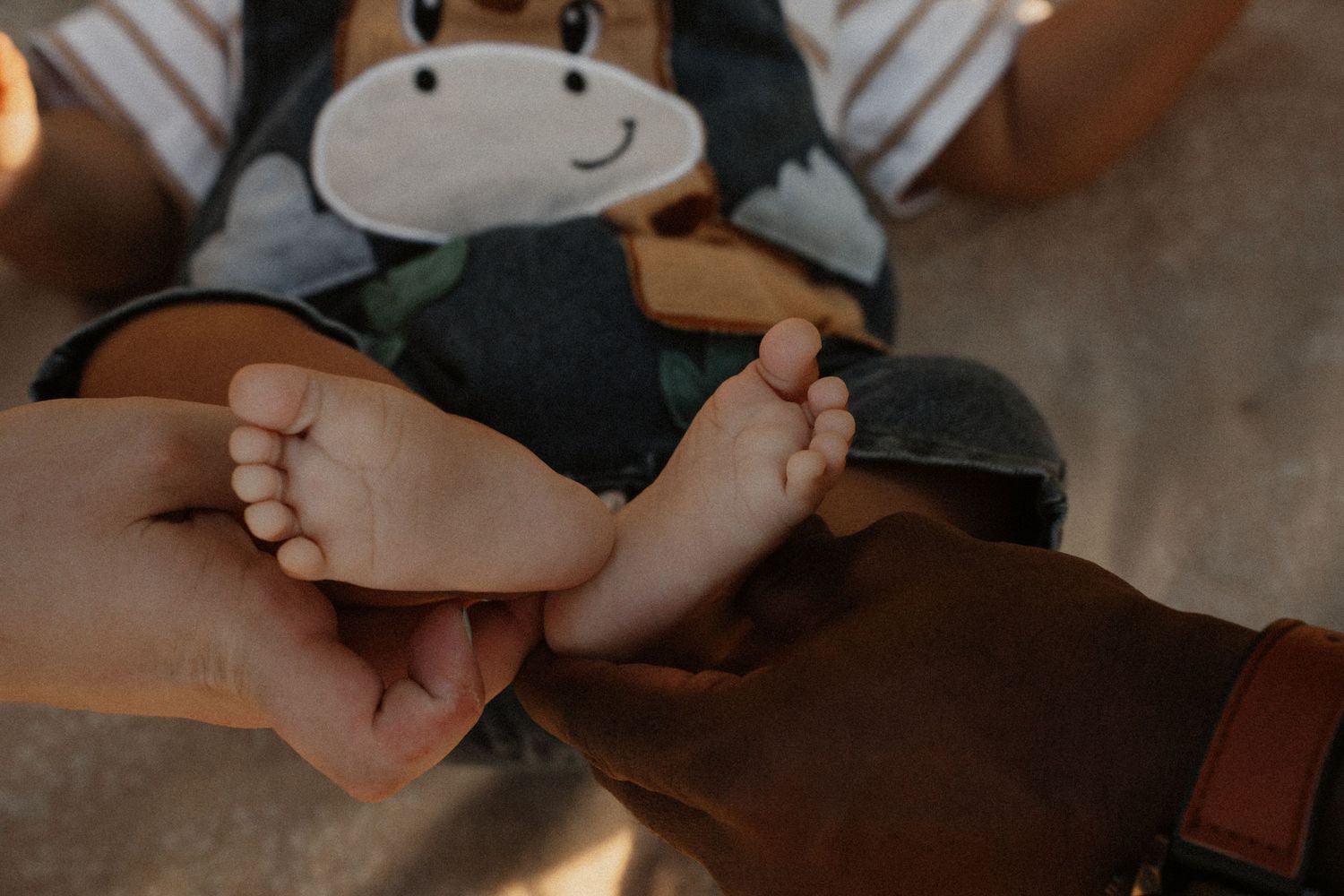 Parents gently holding their newborn baby's tiny feet, baby wearing giraffe outfit on blanket.