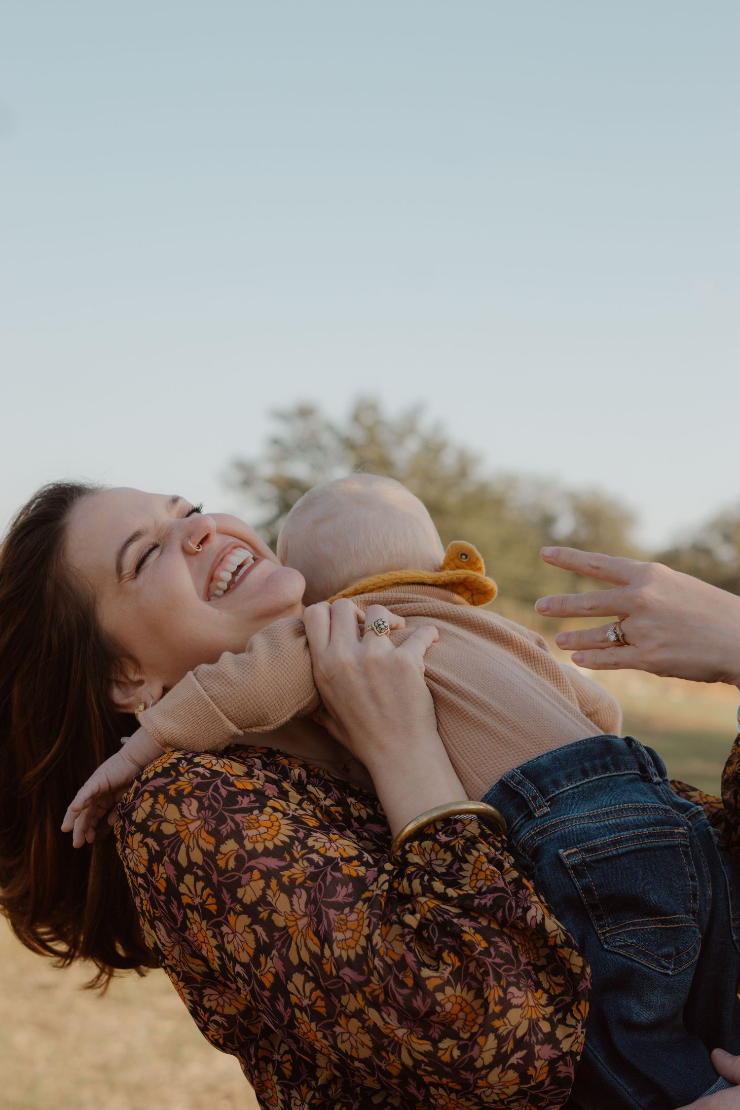 Joyful mother laughs while lifting her baby in golden sunset light during outdoor family session.