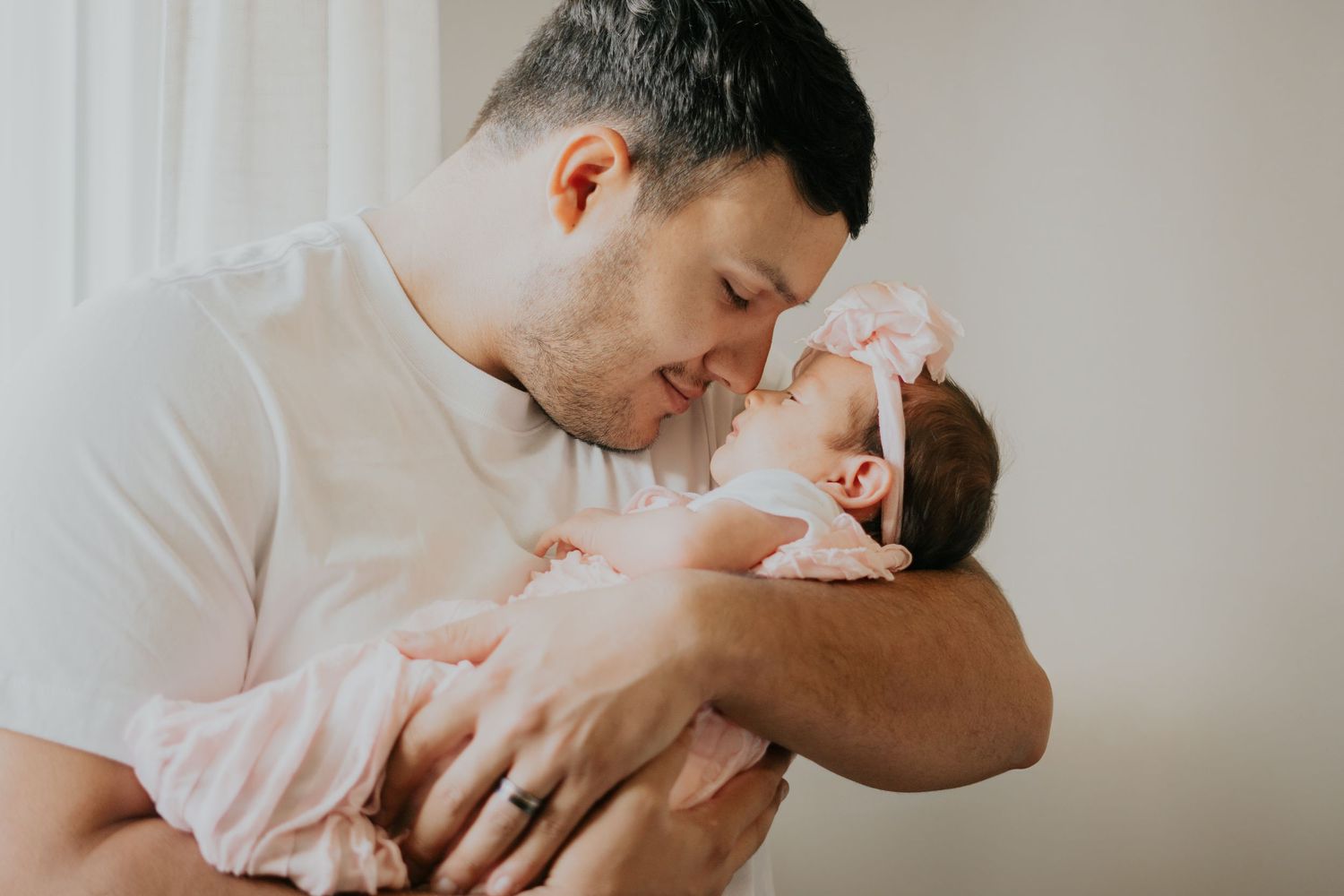 Loving father in white shirt gently nuzzling his newborn baby dressed in pink outfit and bow headband.