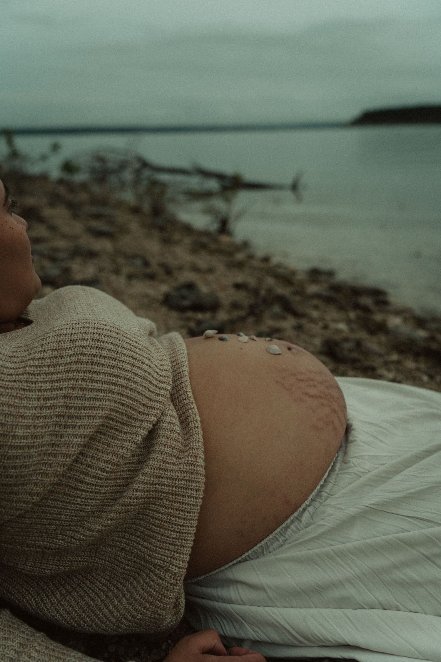A pregnant woman reclines on a rocky beach with small stones placed on her bare bump.