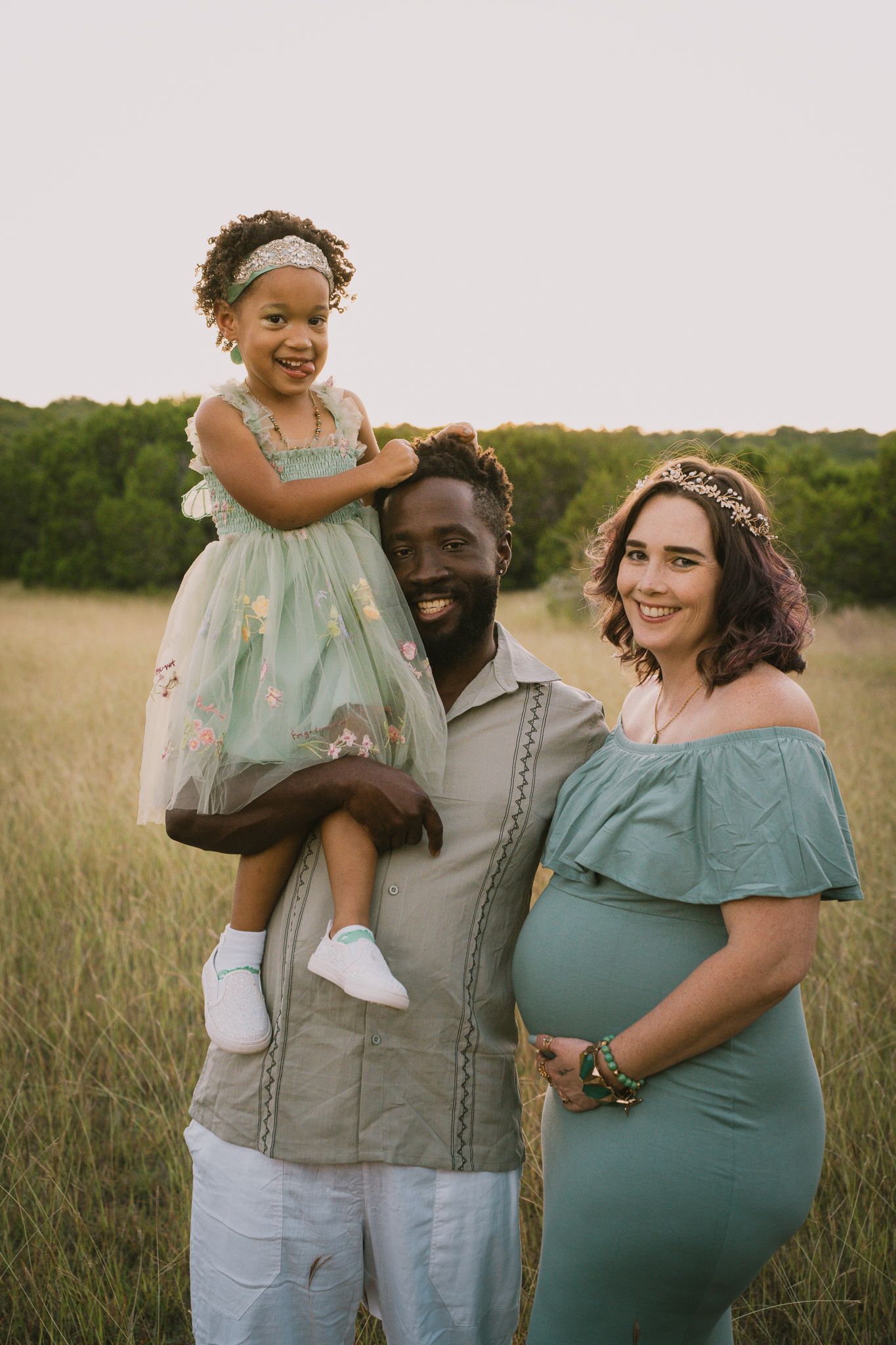 Smiling interracial family with father holding daughter beside pregnant mother in green field.