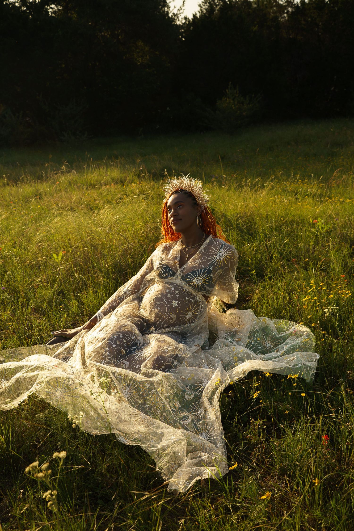 Pregnant woman in a white lace dress sitting in a sunlit green meadow at golden hour.