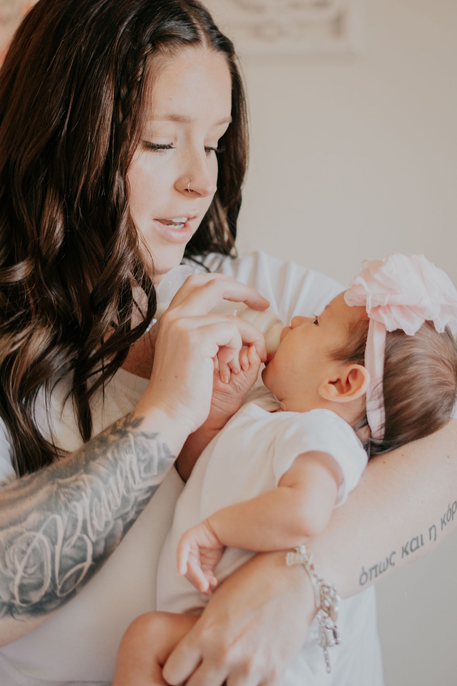 Tattooed mother with long dark hair tenderly holding her newborn baby in a pink bow headband.