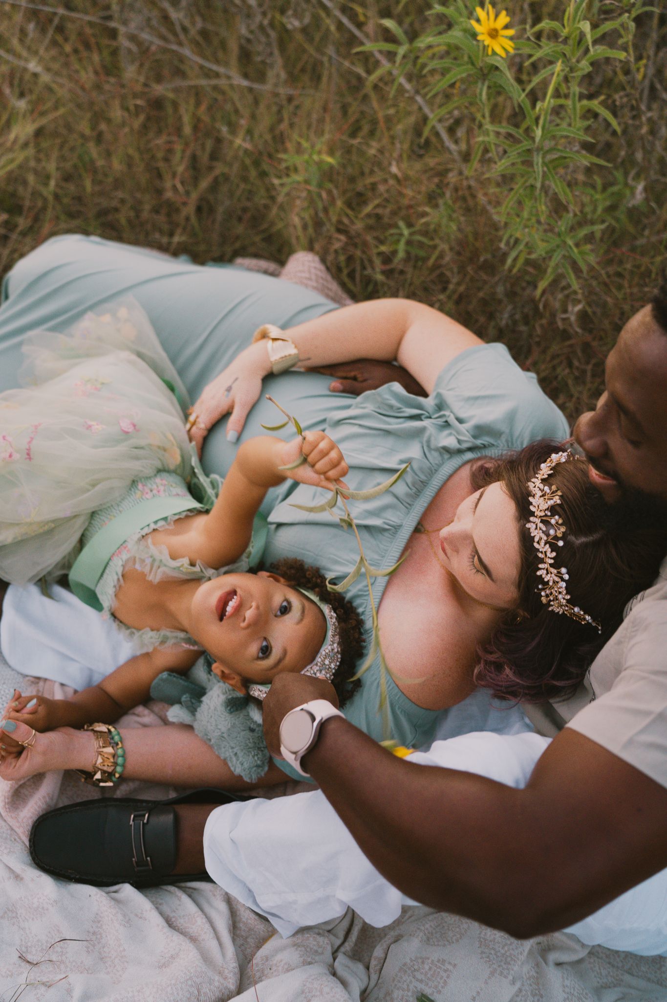 Interracial family lying in meadow with pregnant mother and young daughter smiling joyfully.