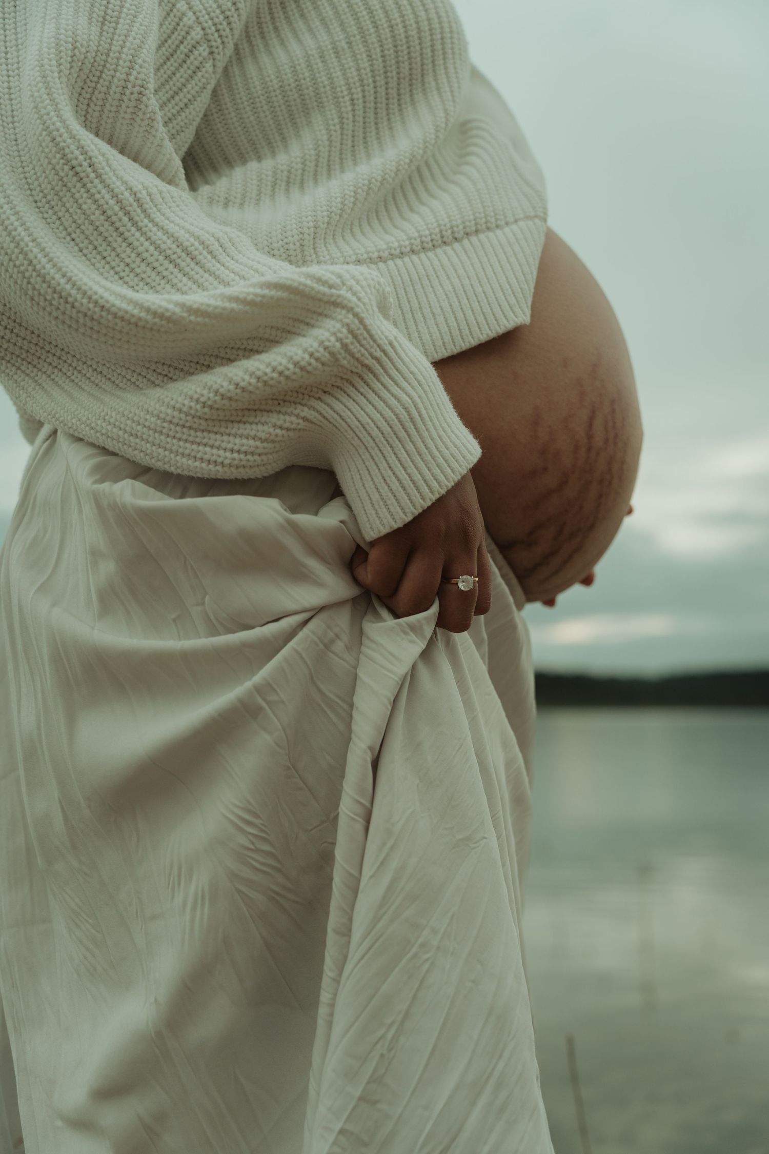 A close-up of a pregnant woman in a white skirt and knit sweater holding her baby bump.