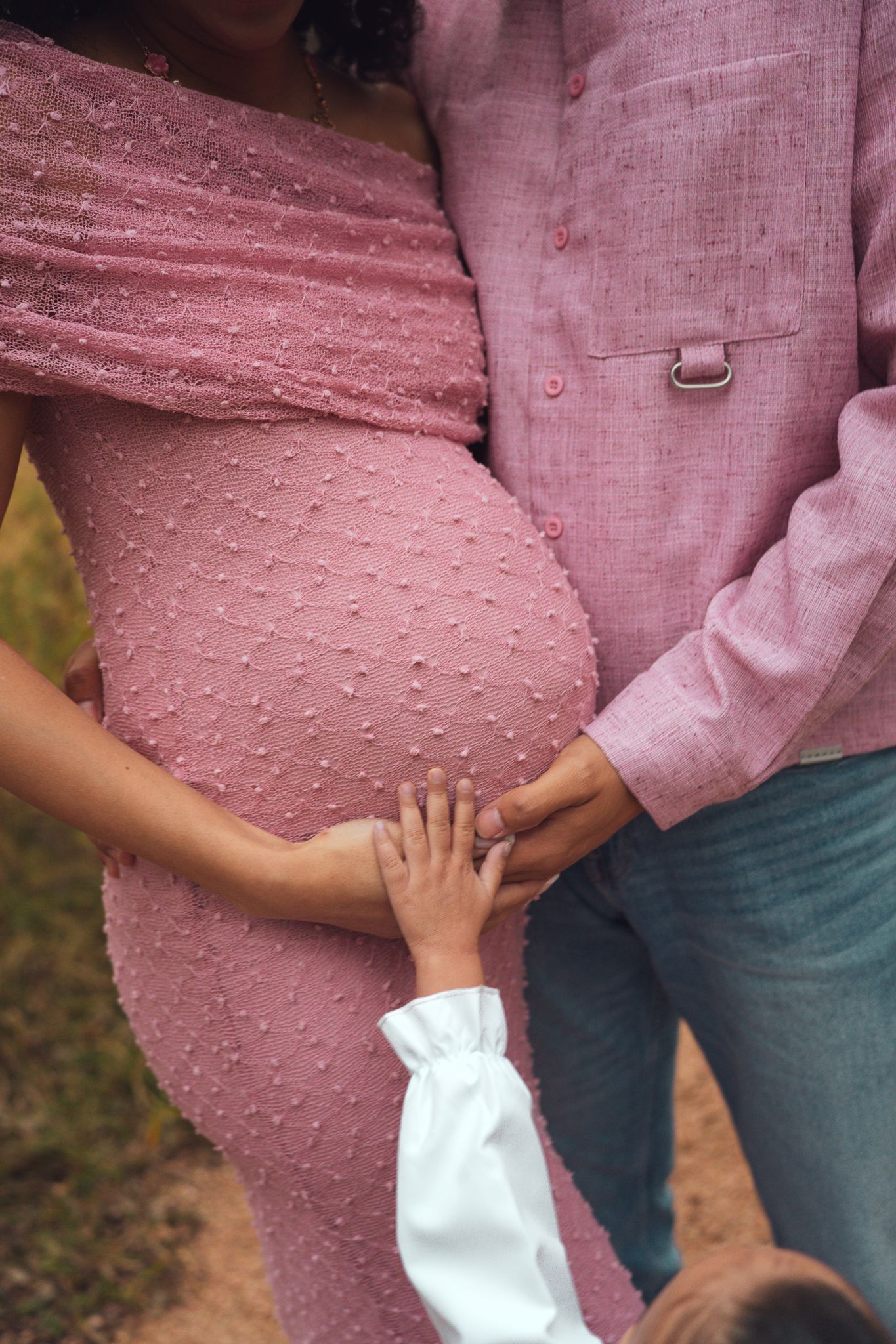 A child places their hand on a pregnant mother's pink dress belly as parents stand close.