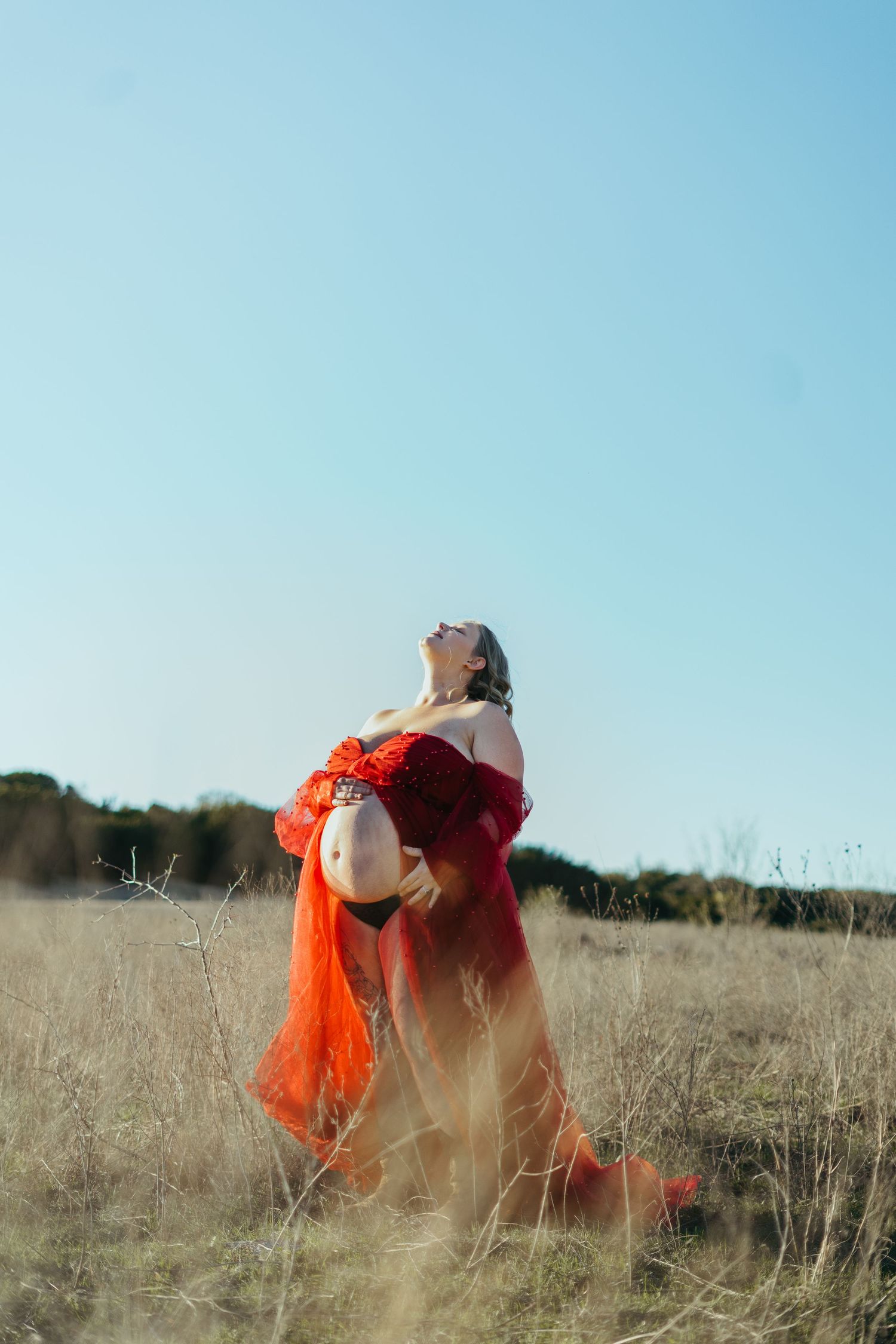 Pregnant woman in flowing red dress stands in dry grass field with head tilted back.