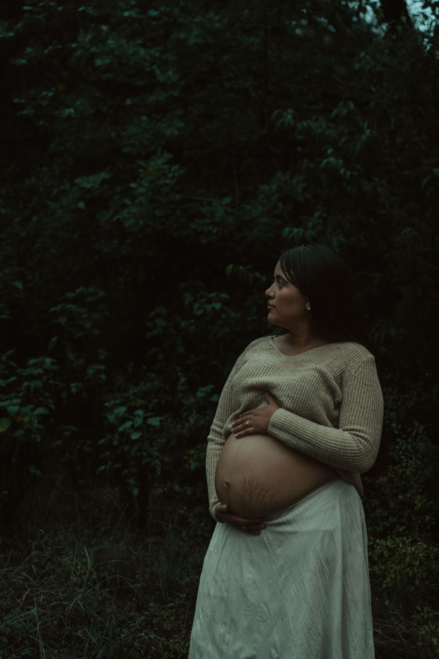 Pregnant woman in beige knit sweater and white skirt holding belly in lush dark forest.