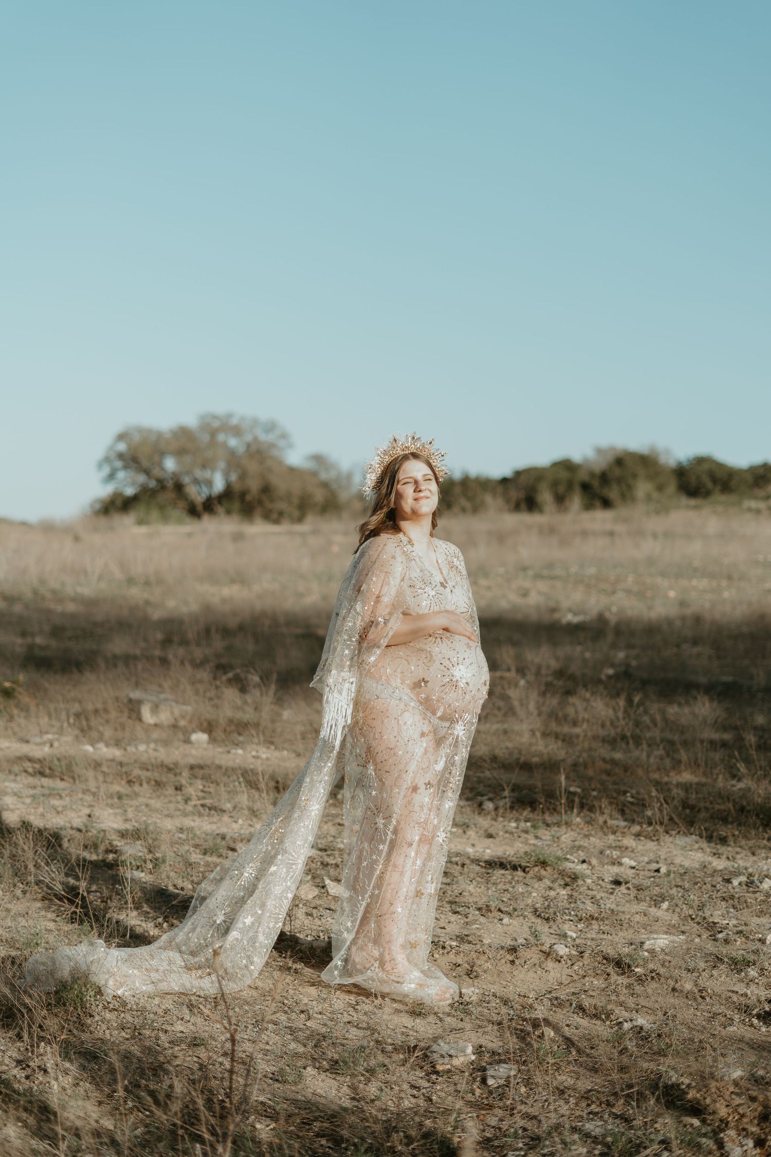 Pregnant woman in sheer lace gown and floral crown standing in golden field under blue sky.