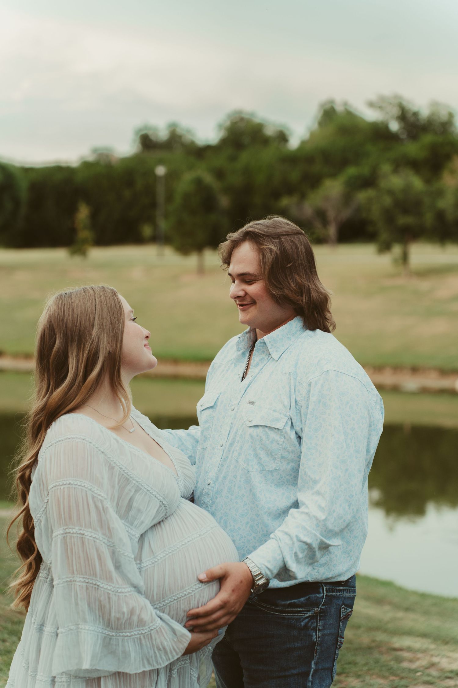 A pregnant couple stands by a pond, smiling at each other during a maternity shoot.