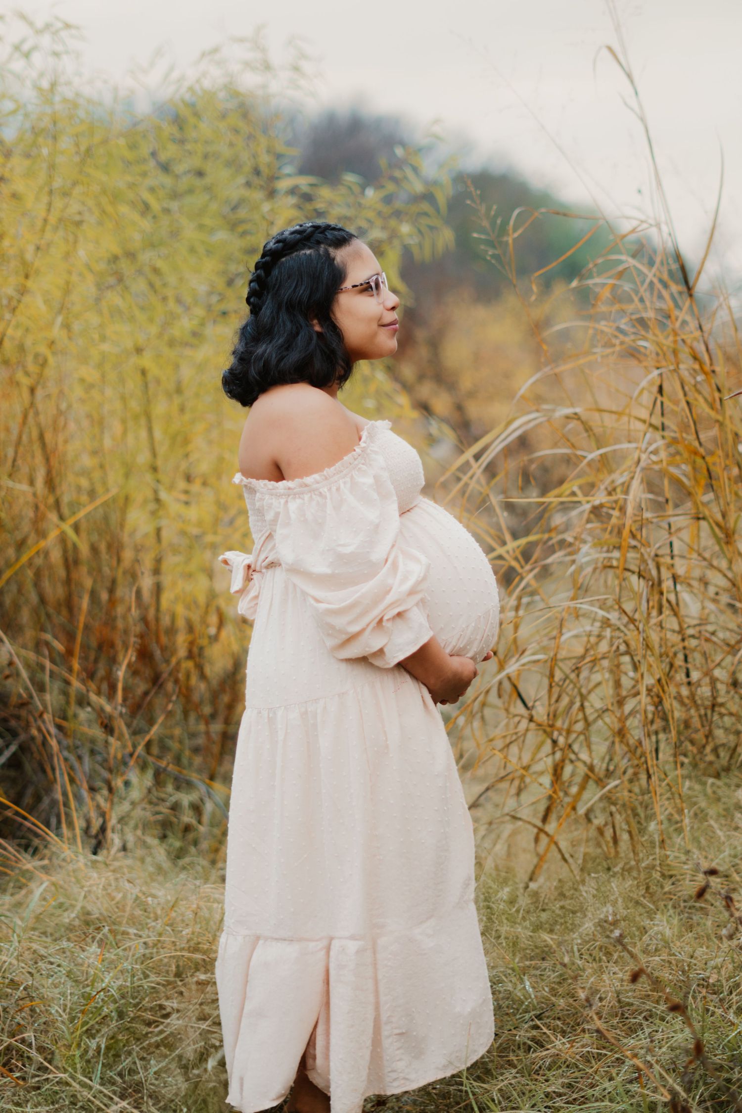 Pregnant woman in off-shoulder white dress cradles baby bump among tall golden grasses outdoors.