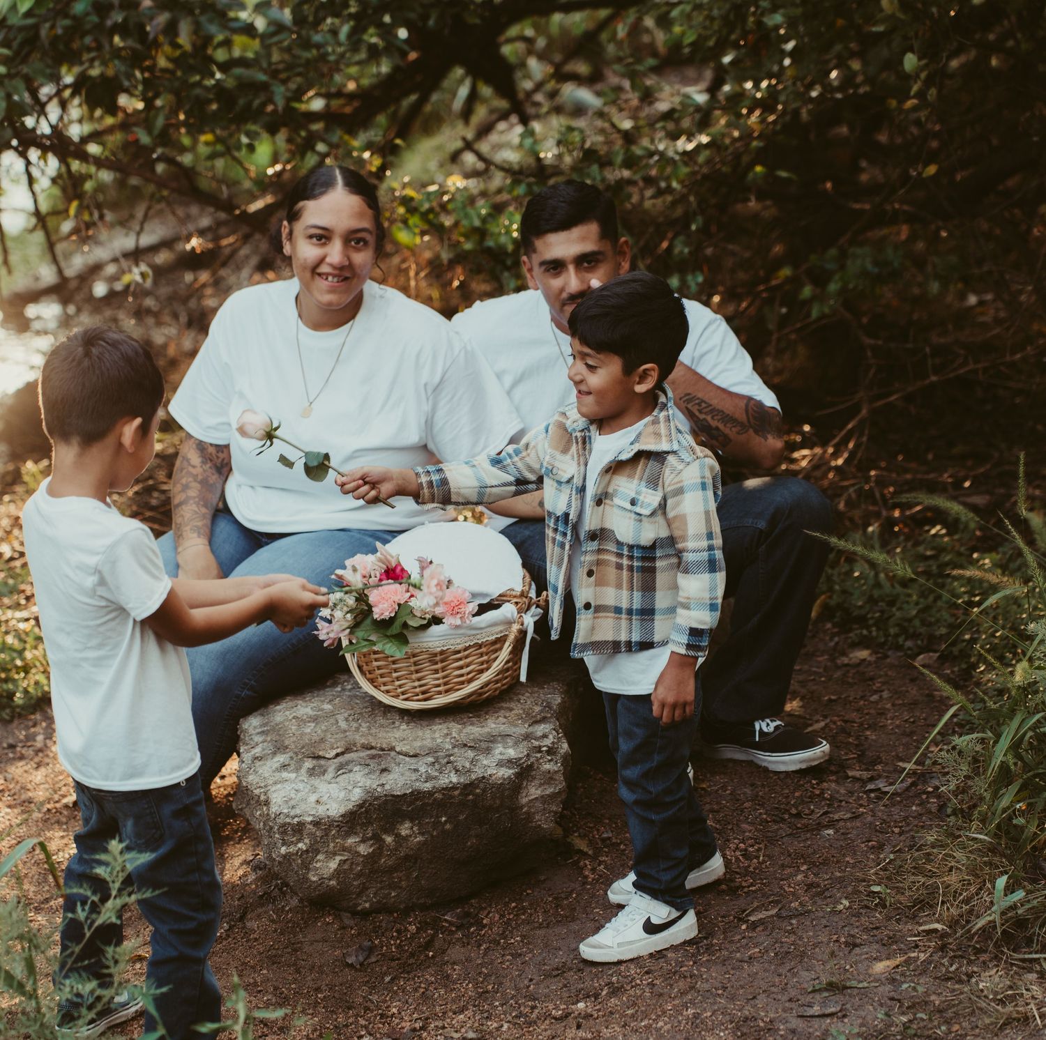 A happy family of four enjoys a picnic with a flower basket in a wooded outdoor setting.