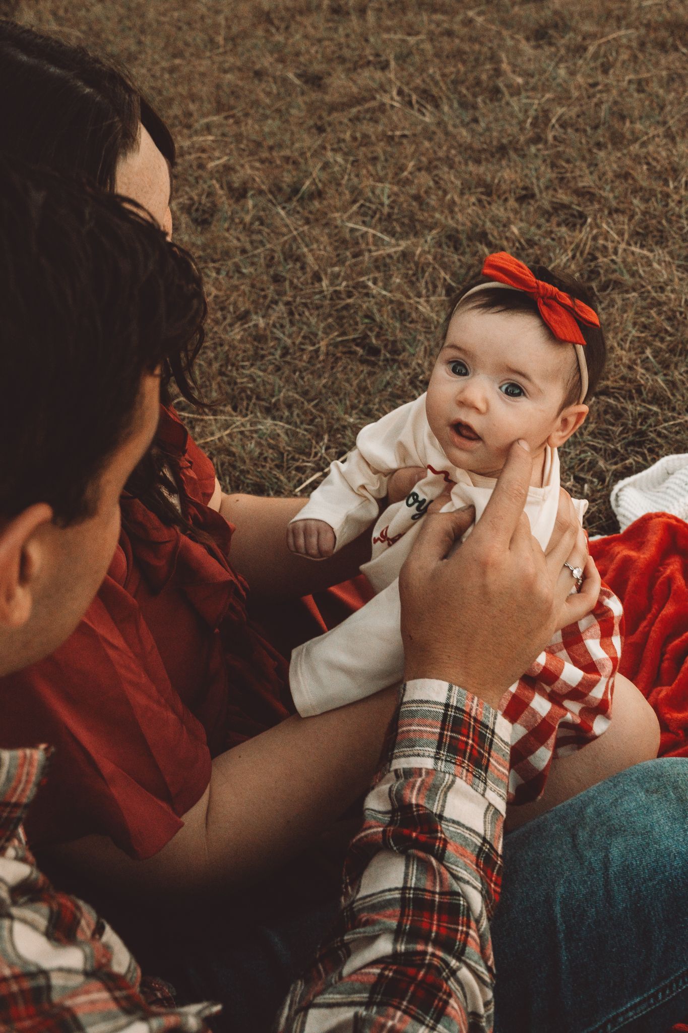 Parents sitting in grass interacting with adorable baby girl wearing red bow headband.