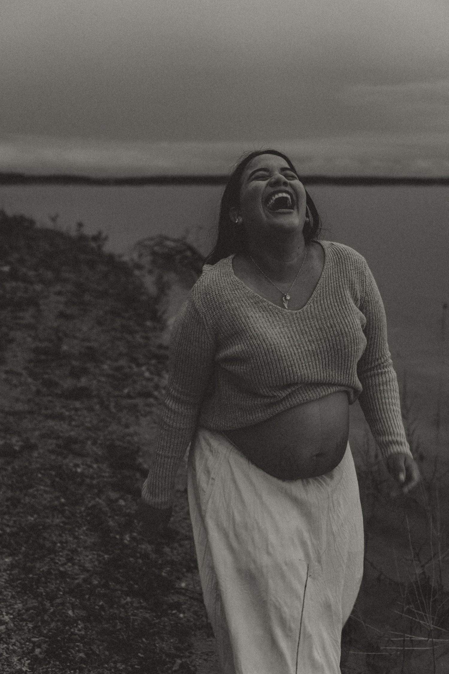 Black and white photo of joyful pregnant woman laughing outdoors near water in knit sweater.