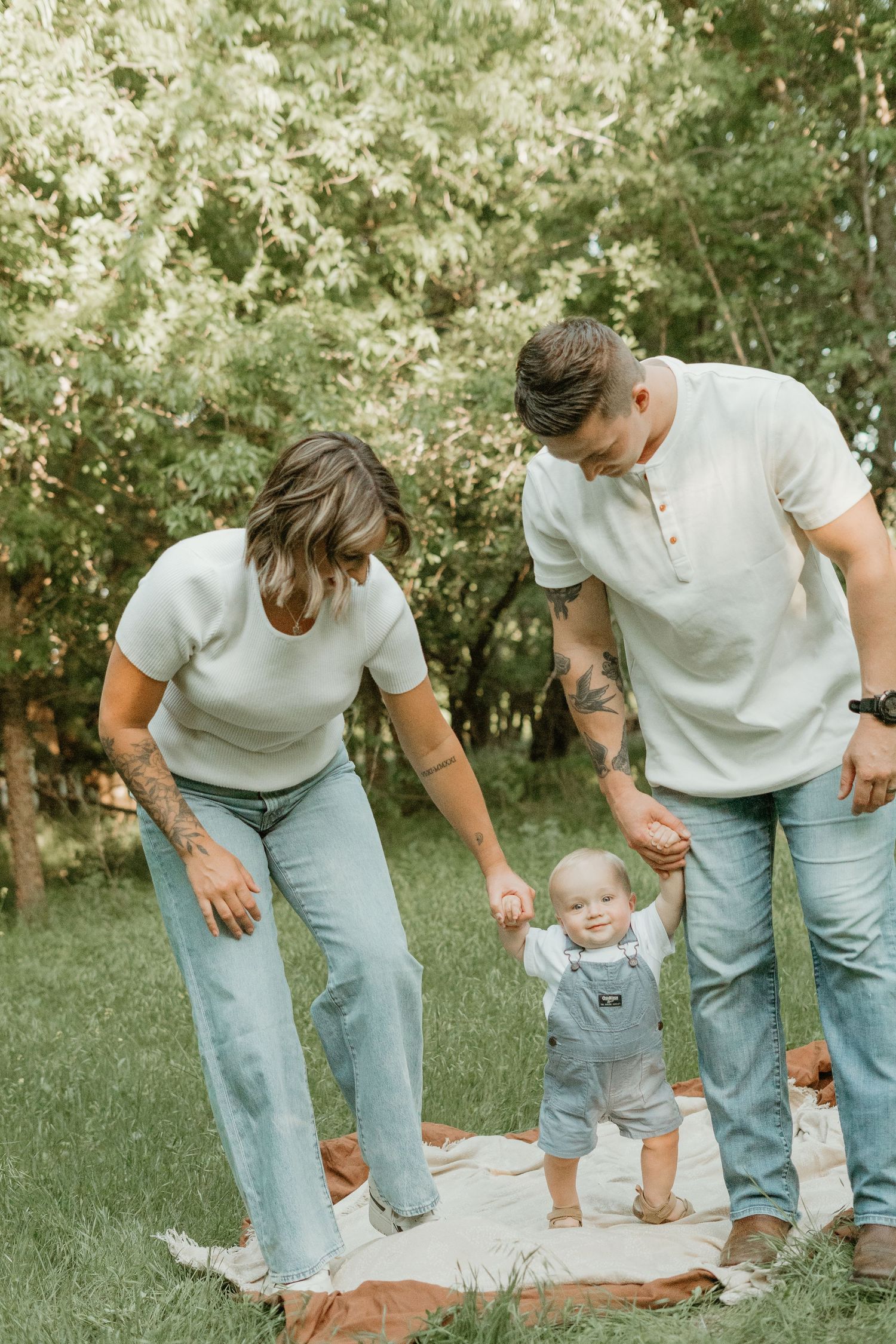 Young family helps baby take first steps on blanket in lush green park during outdoor photo session.