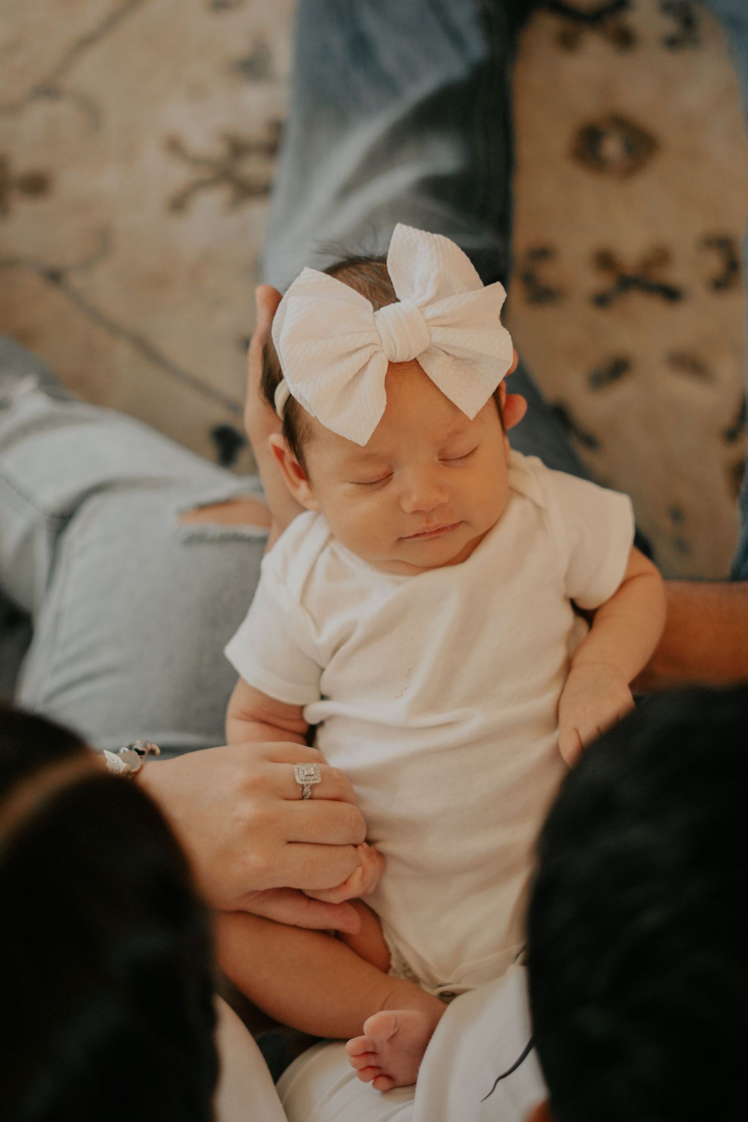 Sleeping newborn baby girl in white onesie and bow headband cradled by parents hands.