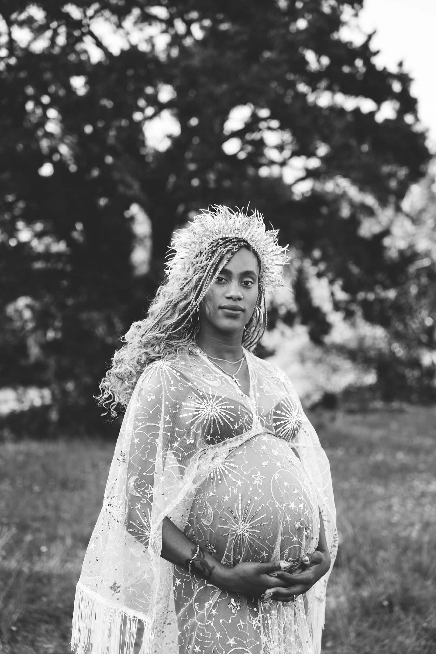 Black and white portrait of a pregnant woman in a lace dress and floral crown outdoors.