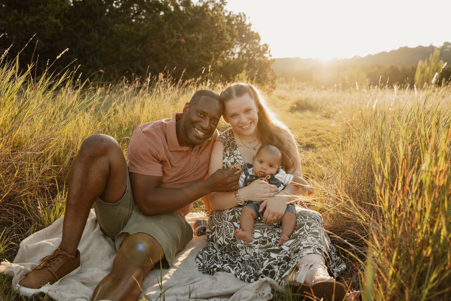 Smiling interracial family sitting together in golden sunlit field, parents with baby at sunset.