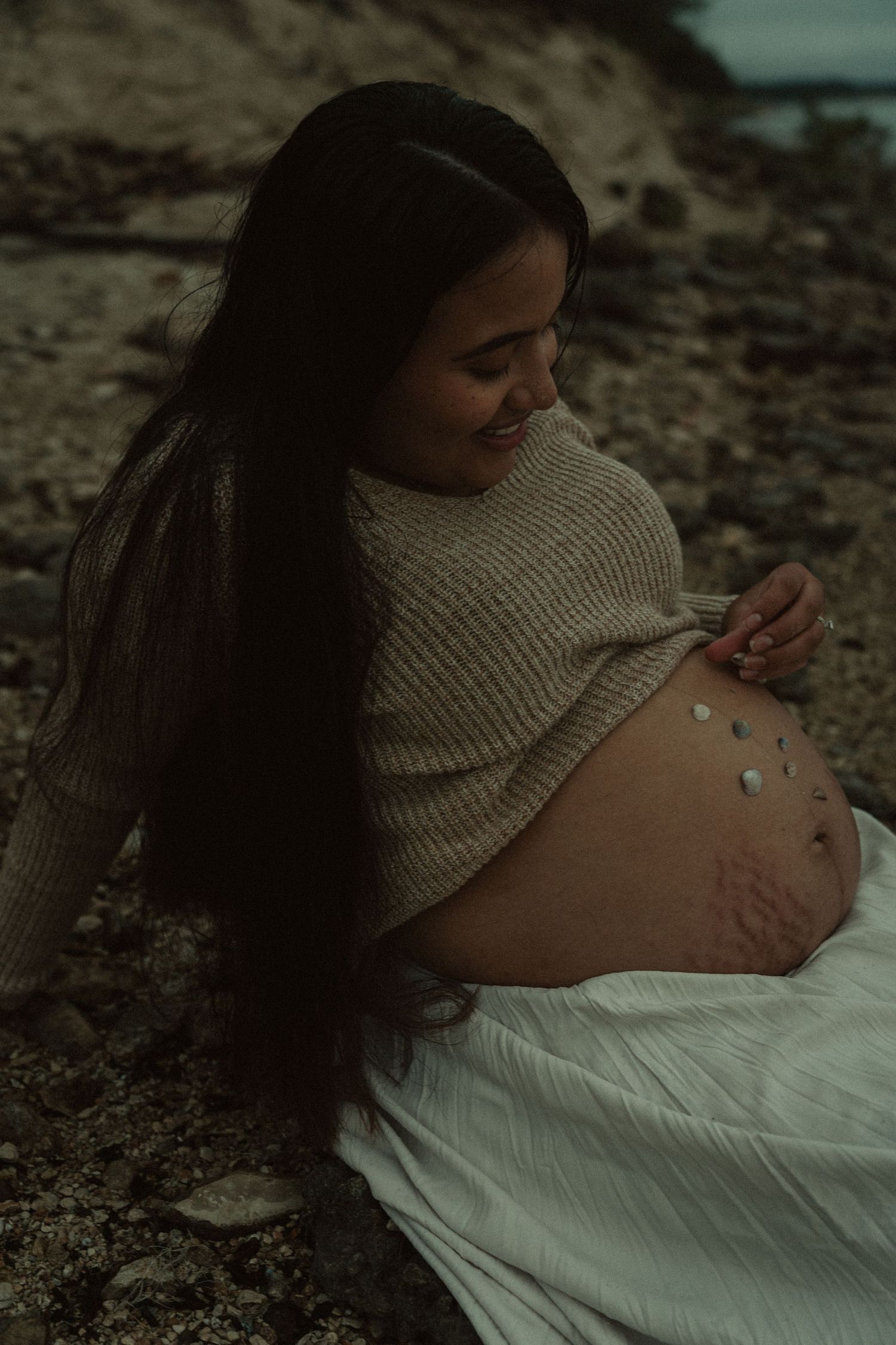 A smiling pregnant woman in a knit sweater sits on a pebble beach with stones on her bump.