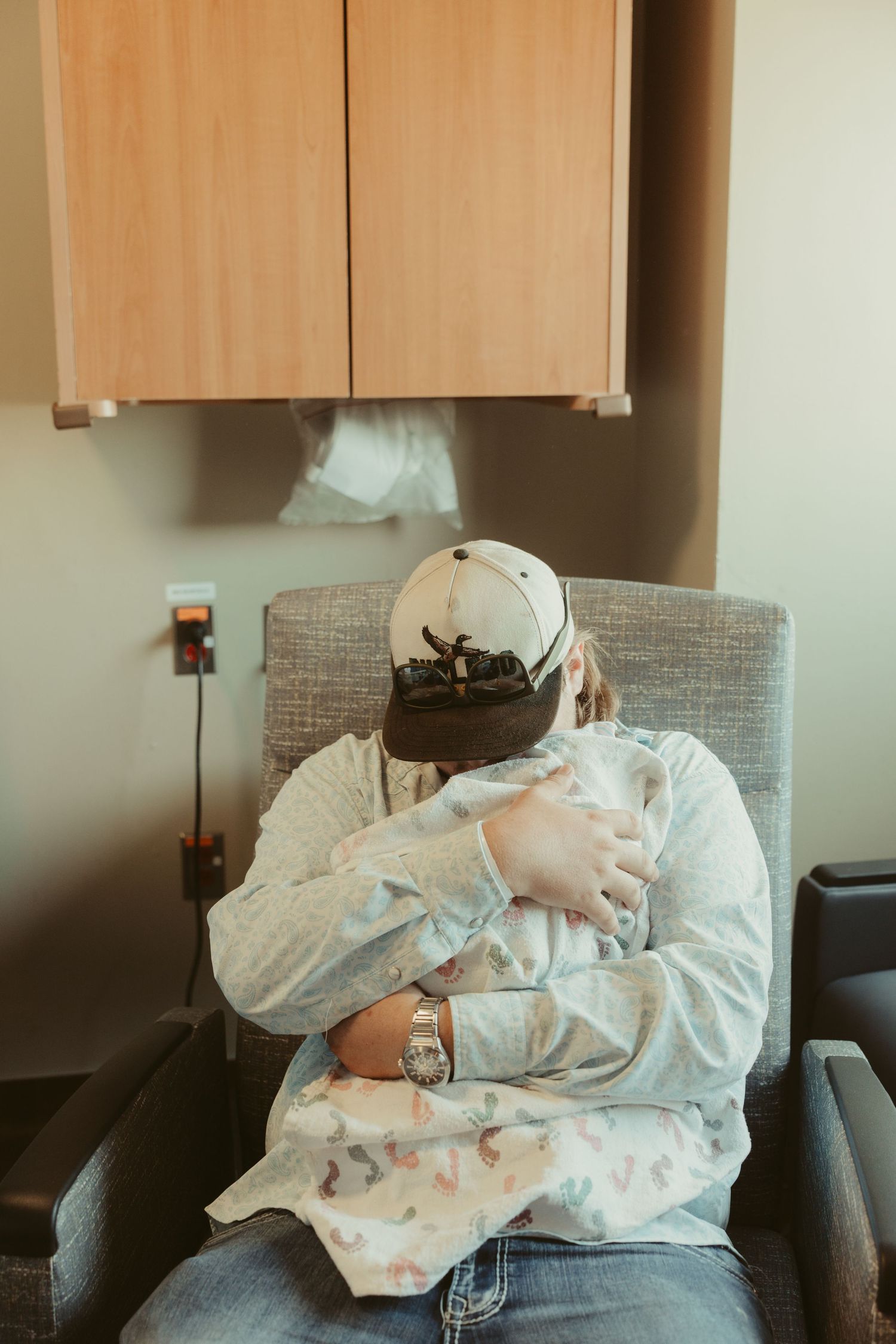 A new father sits in a hospital chair cradling his newborn baby.