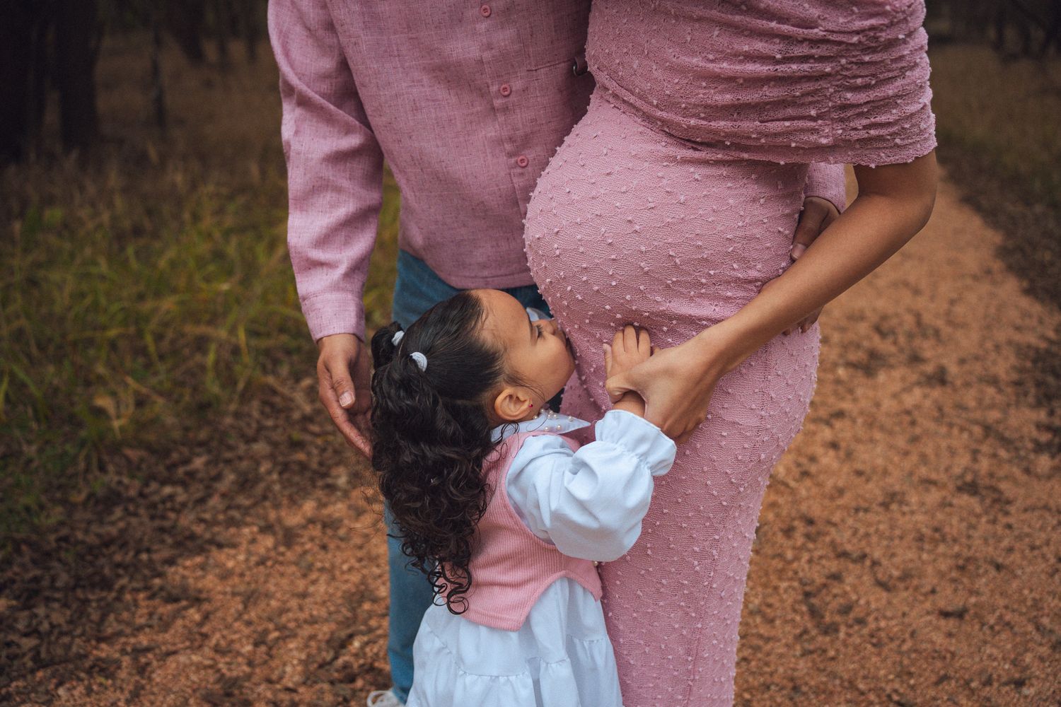 Young girl in white dress hugging pregnant mother's belly outdoors in pink maternity gown.