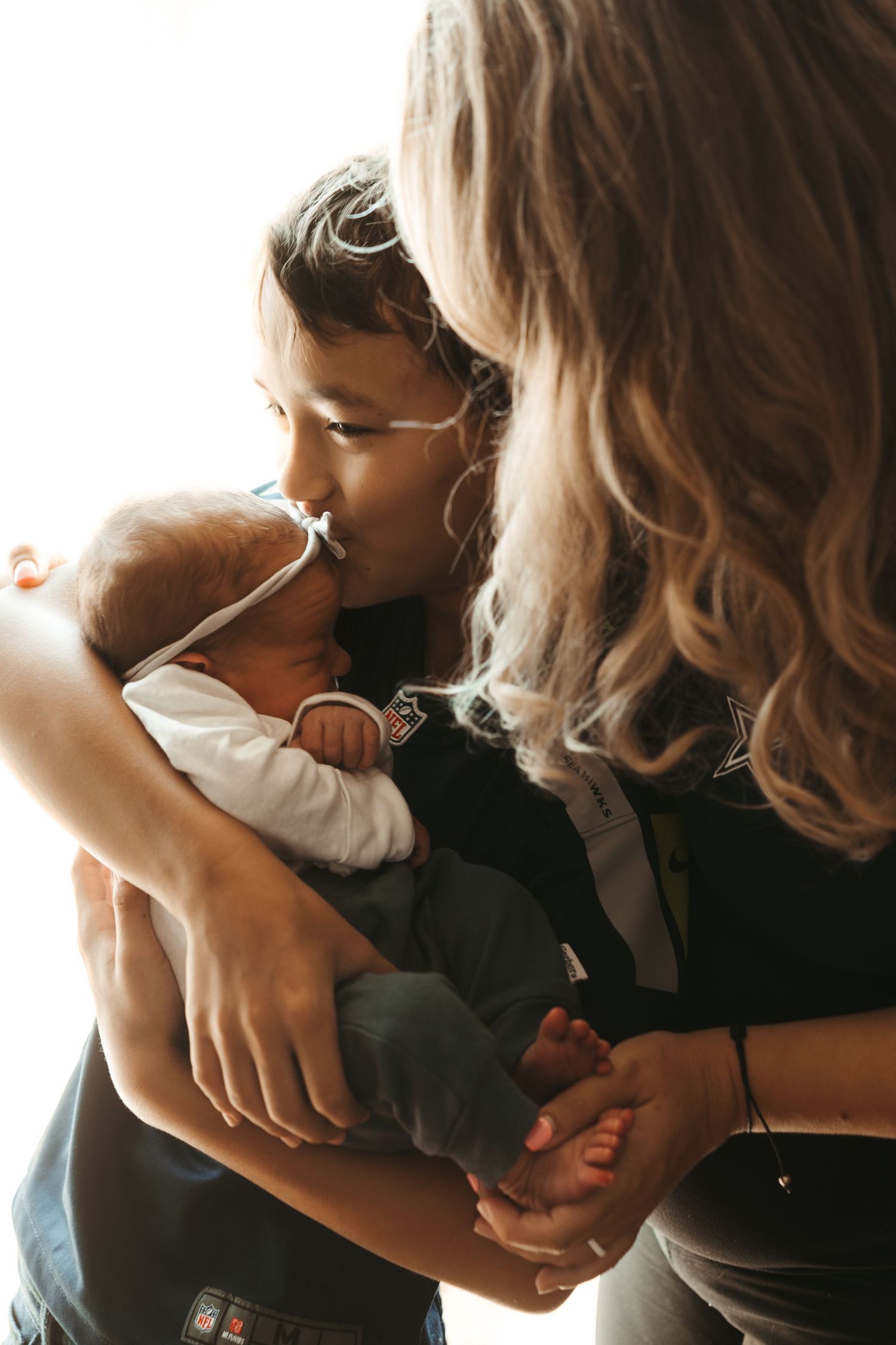 A young child kisses a newborn baby while a blonde woman looks on tenderly.
