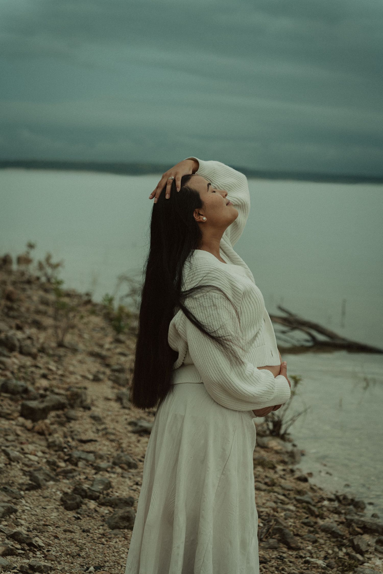 A pregnant woman in white places her hand on her head while standing by a calm lake.