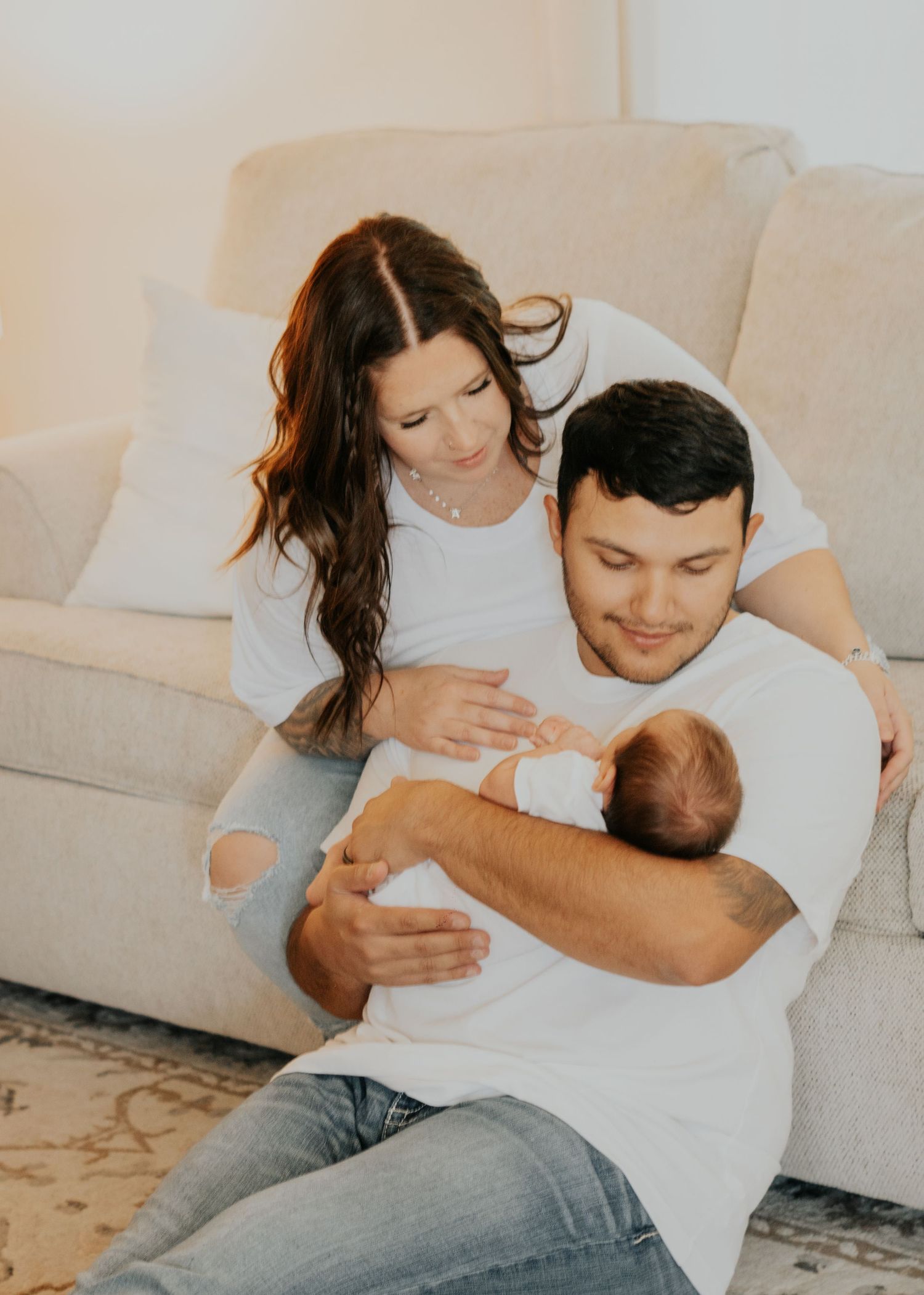 Young couple in white shirts sitting on floor together lovingly holding their newborn baby.