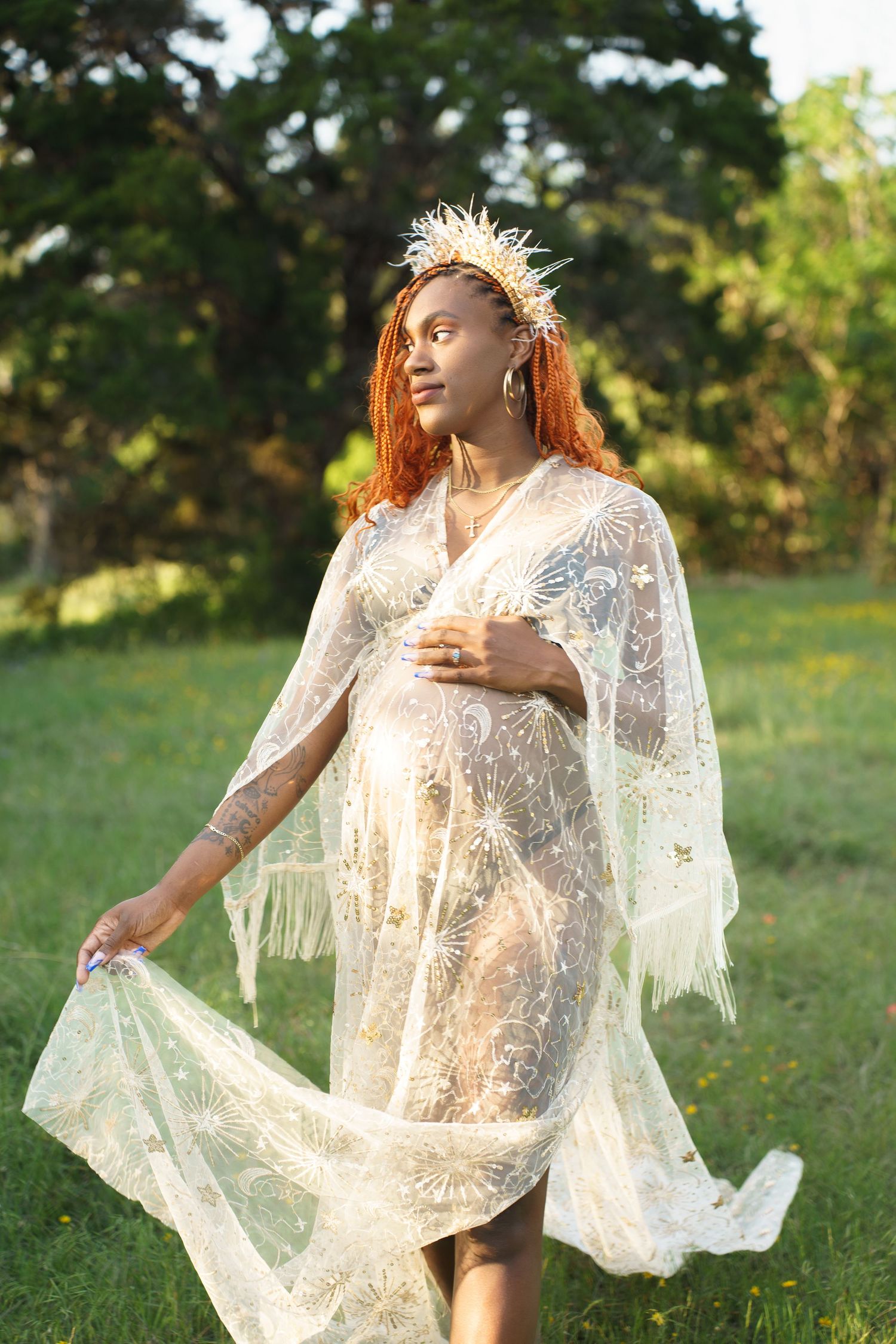 Pregnant woman with orange braids wearing a celestial lace dress and crown in a sunny field.