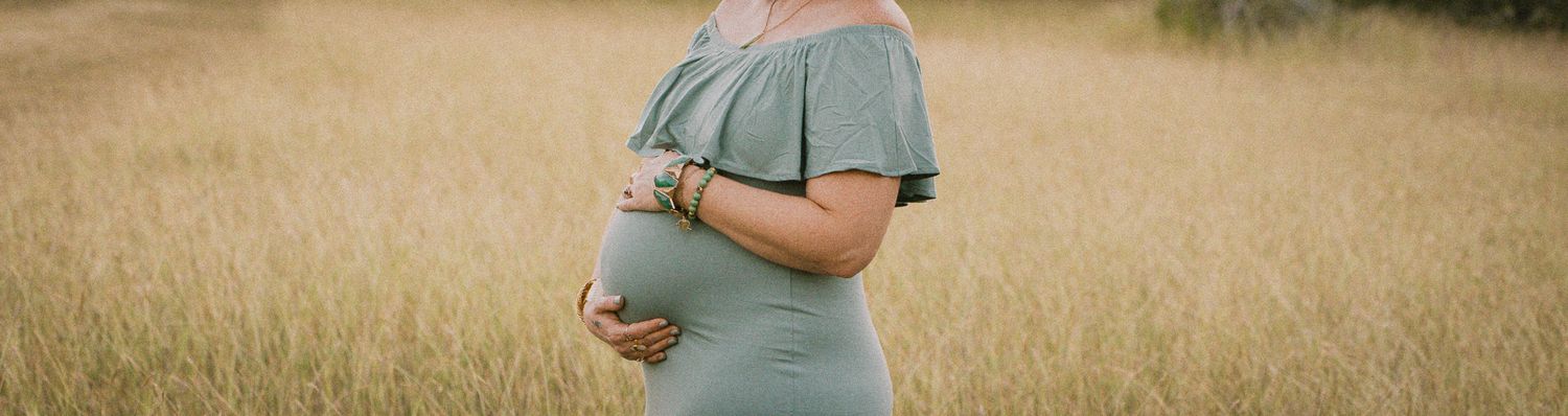Pregnant woman in sage green off-shoulder maternity dress standing in golden wheat field.