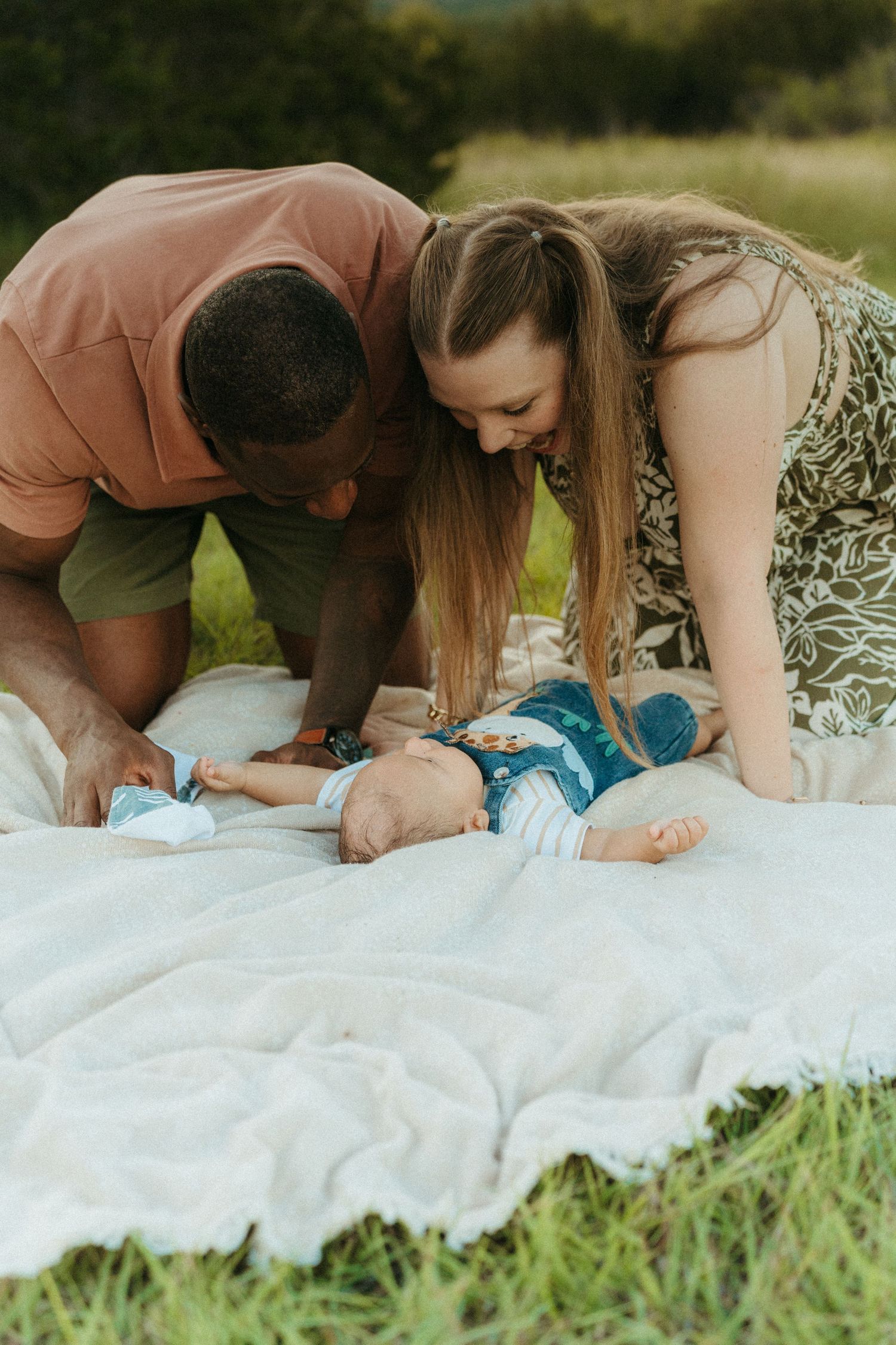 Happy interracial couple leaning over their baby lying on a white blanket outdoors.
