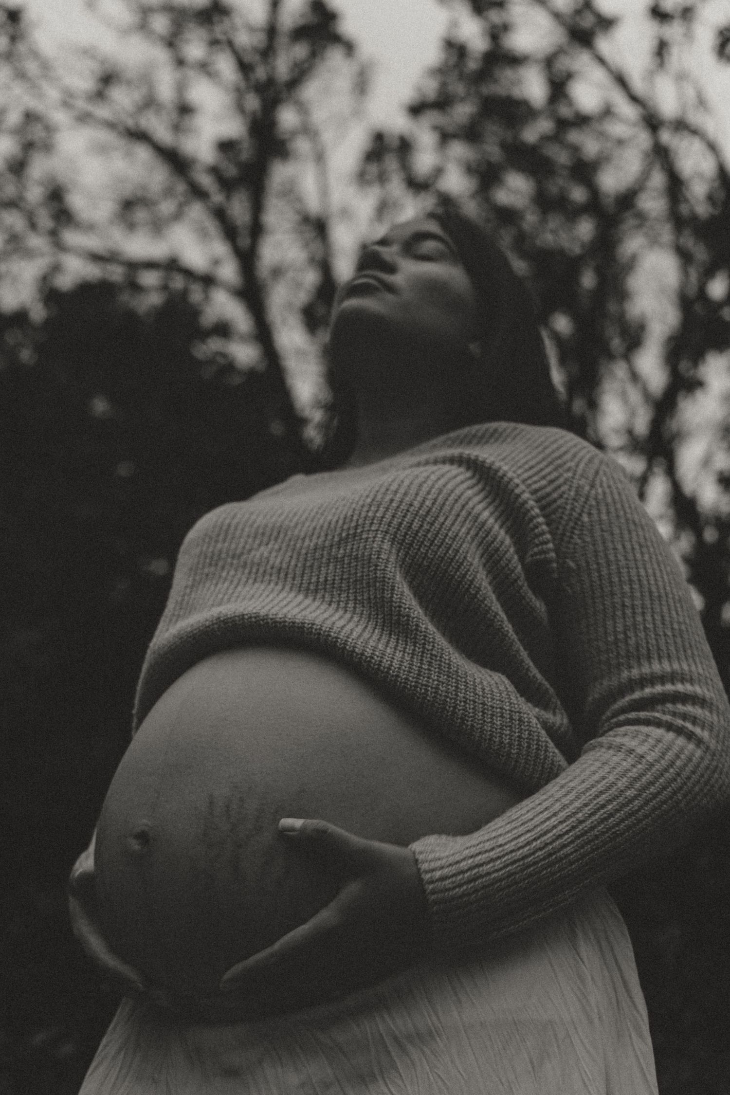 Black and white artistic photo of pregnant woman cradling belly with trees in background.