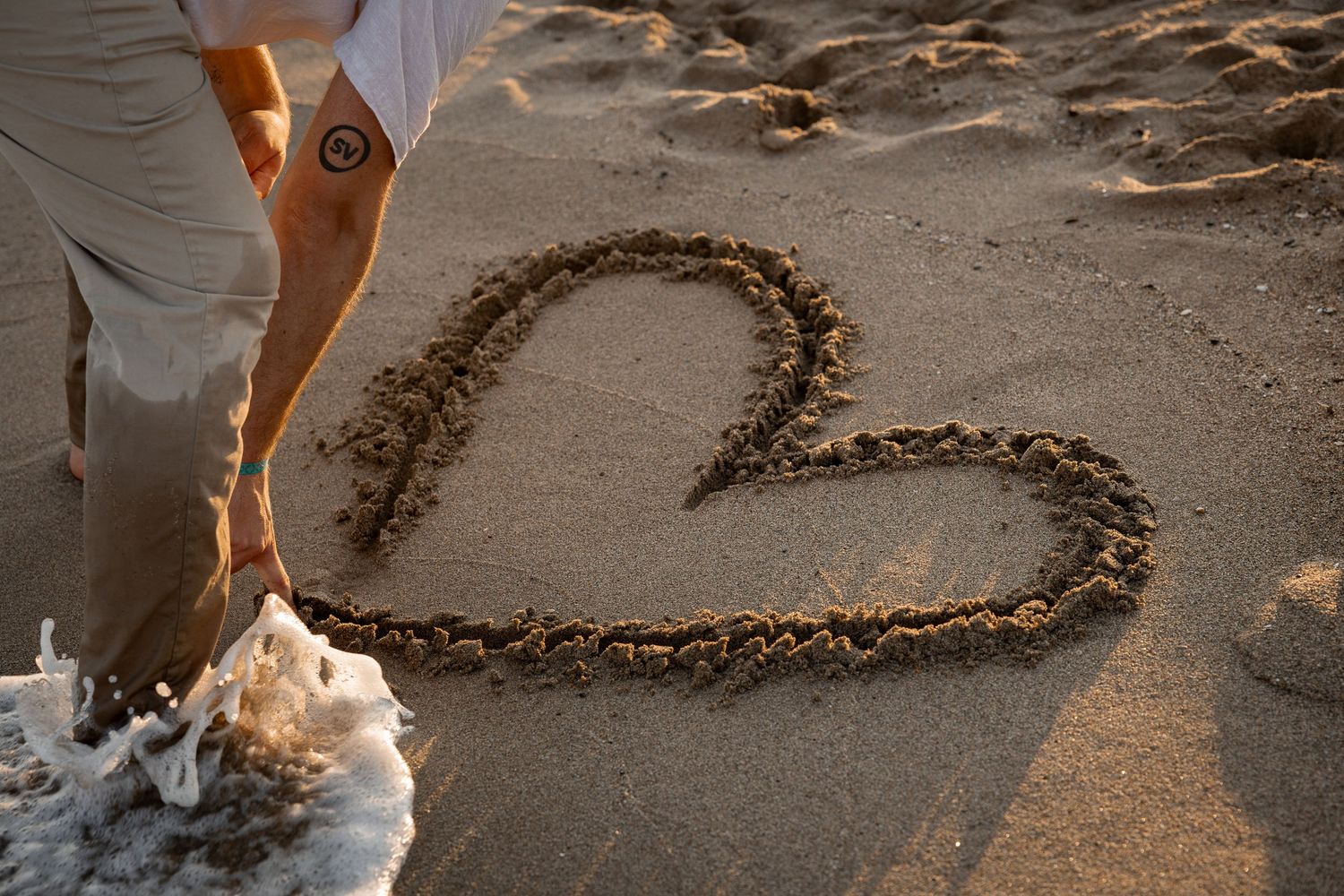 Homme dessinant une forme de cœur dans le sable humide de la plage au coucher du soleil avec les vagues de l'océan dans l'Hérault à Béziers.
