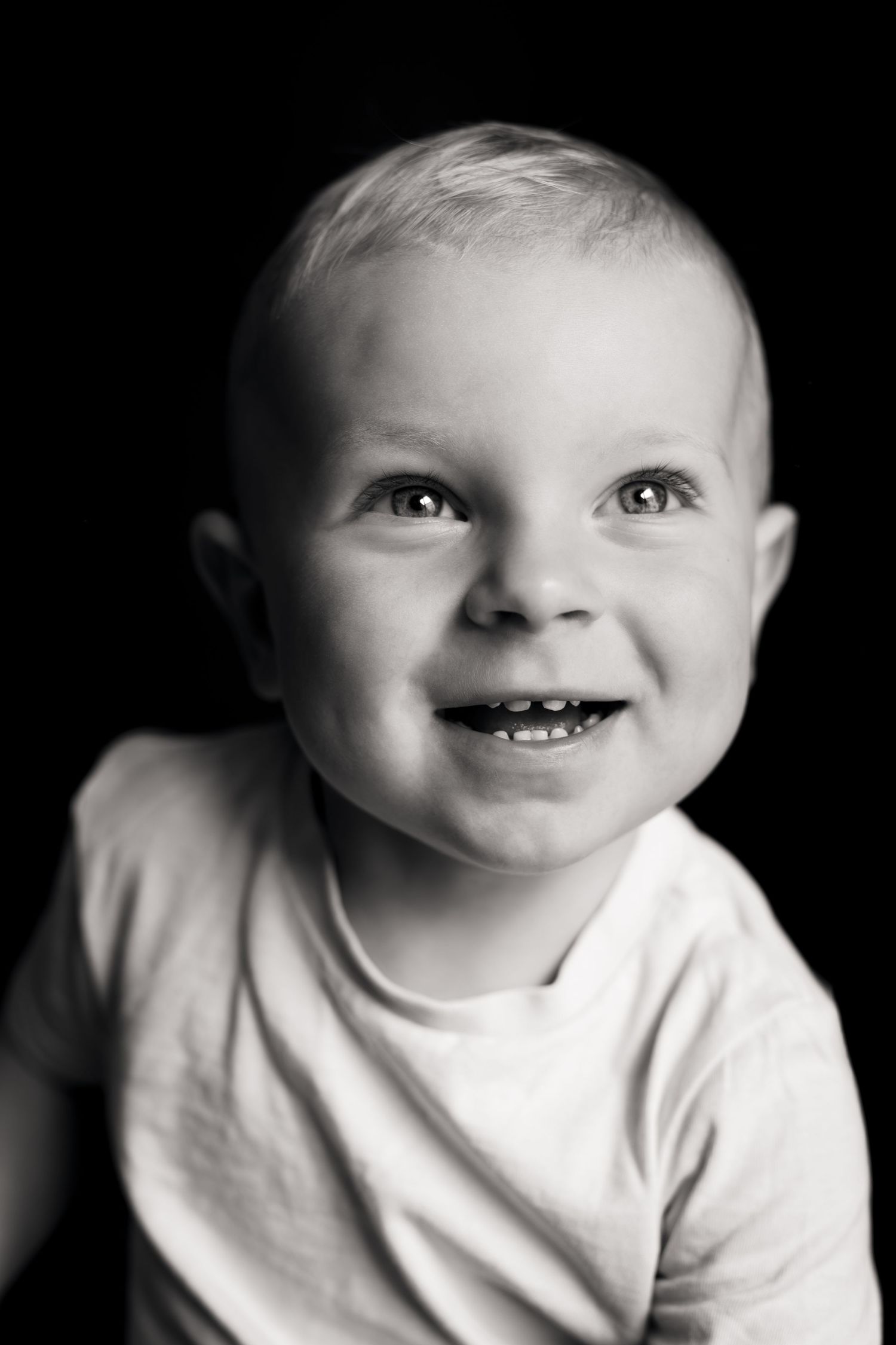 Black and white portrait of a smiling baby with tiny teeth, wearing a white shirt against a dark background.